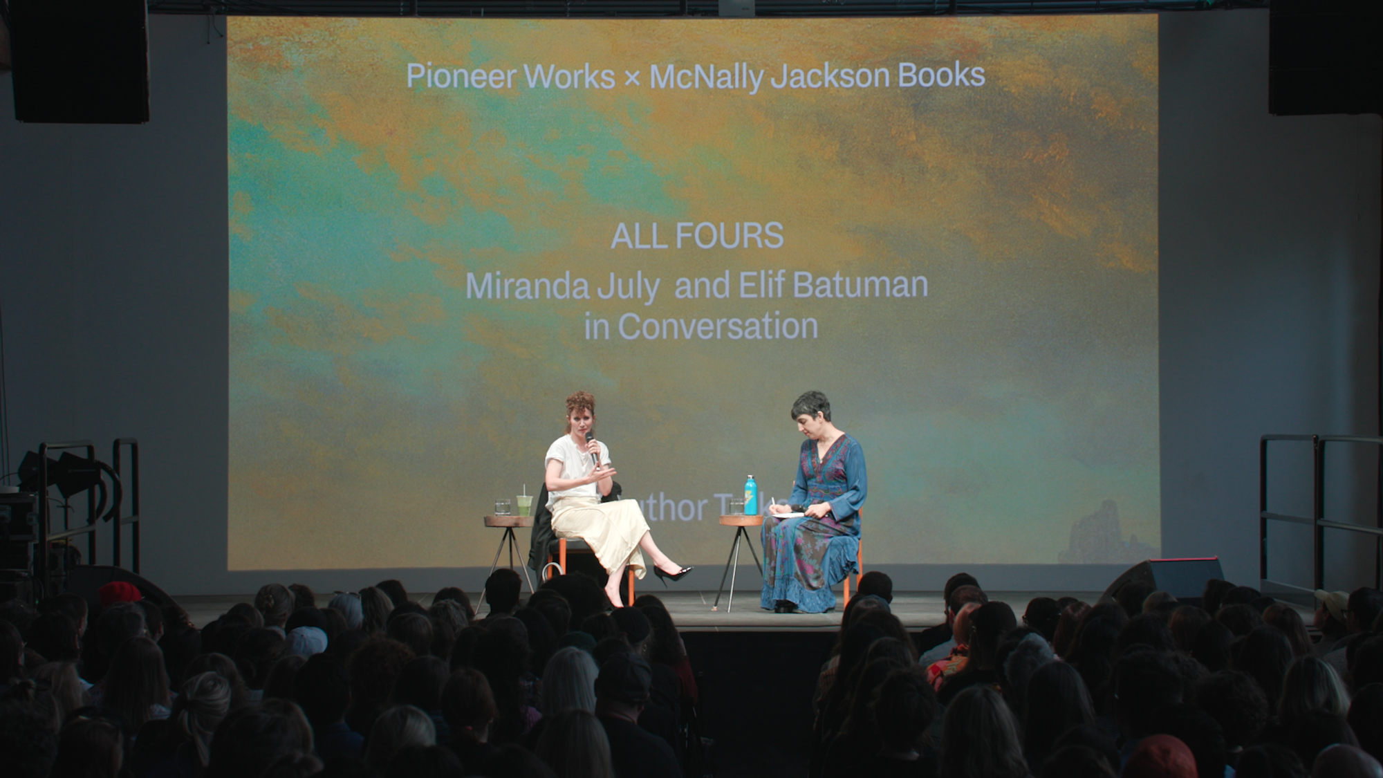 Miranda July and Elif Batuman sit on a spotlit stage, speaking into microphones for an enraptured crowd.