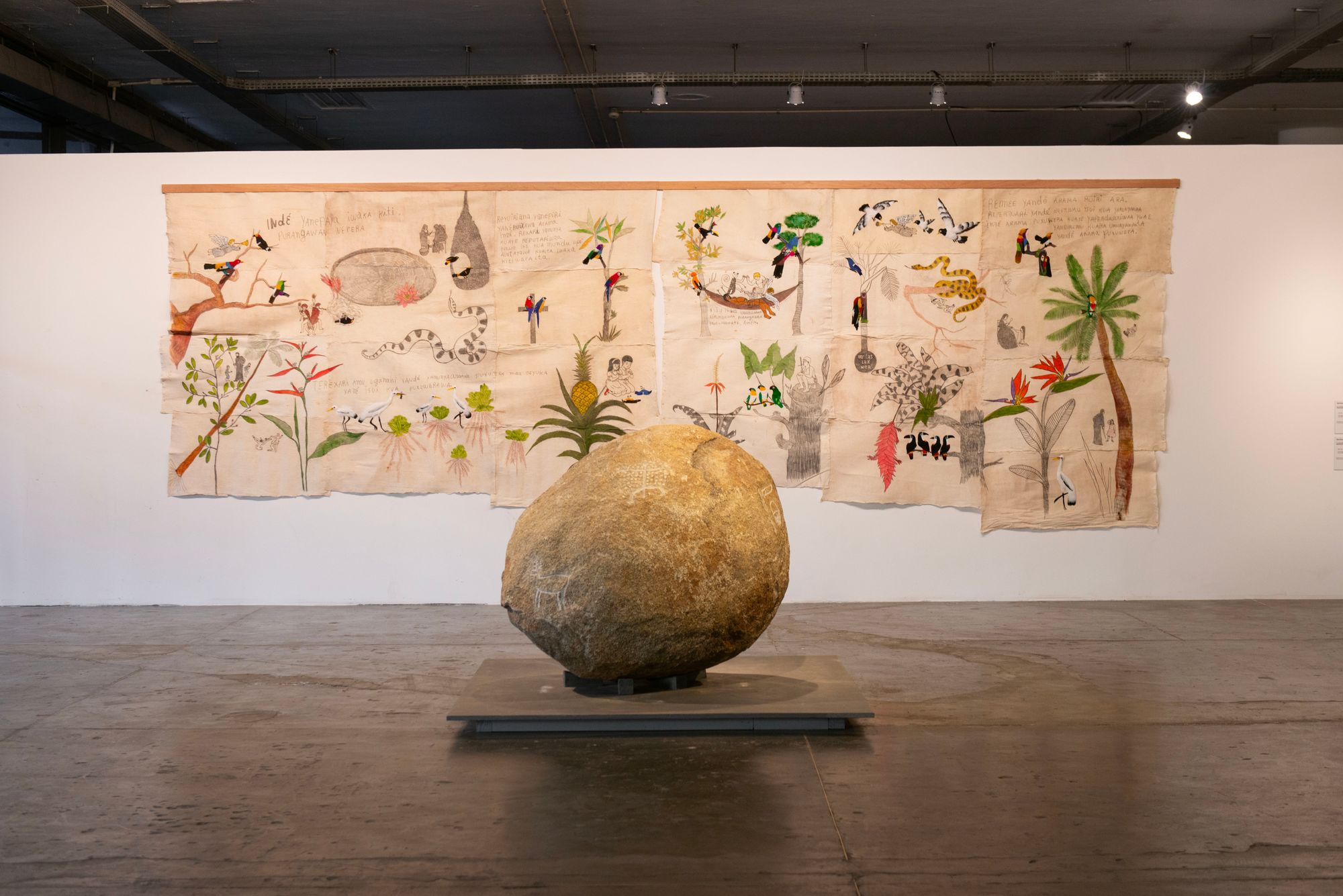 A color photograph of an exhibition space where a large rock rests on a platform in front of a white wall covered in some sort of quilt. 