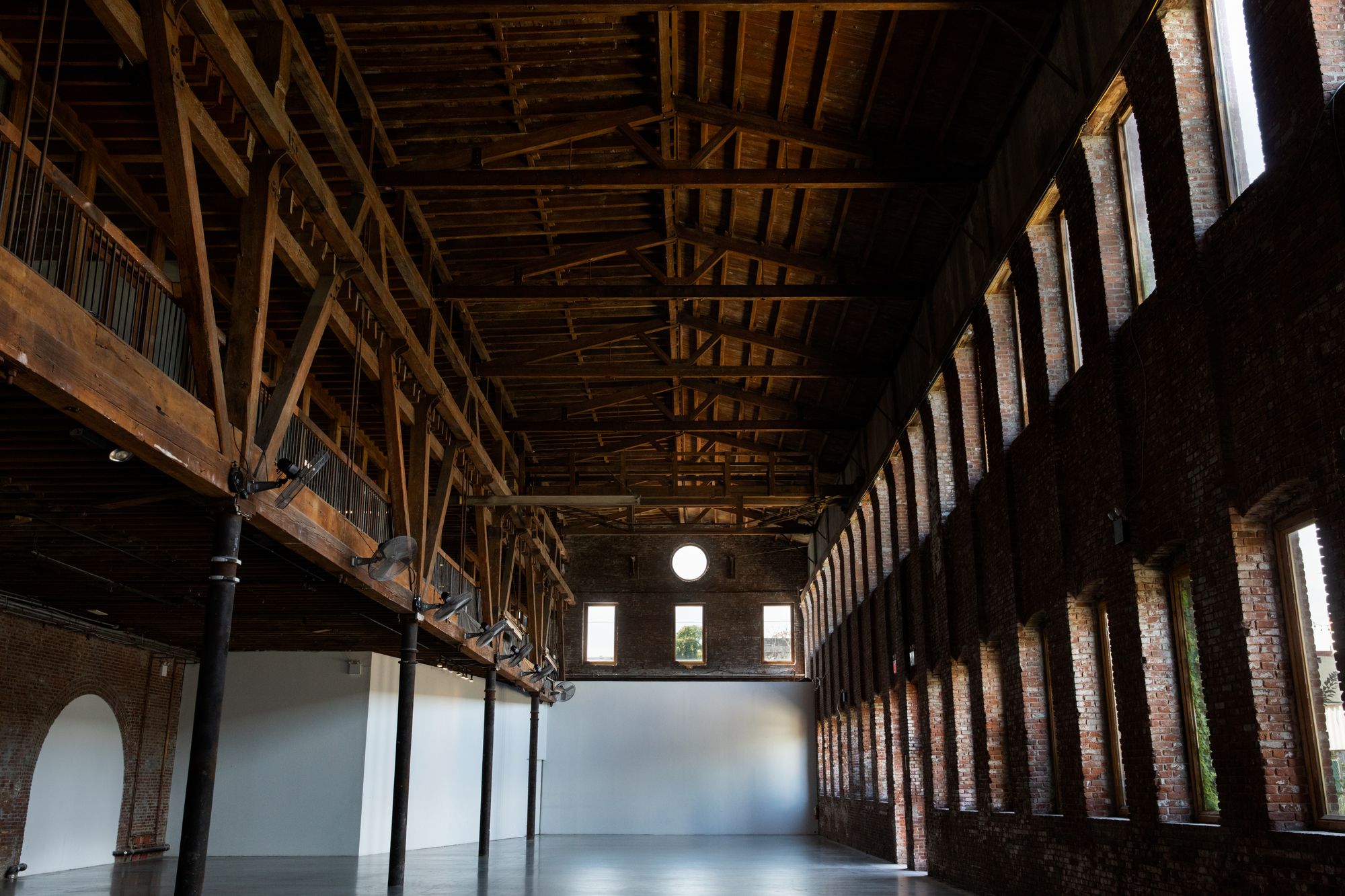 A large empty room, with brick walls, a wooden mezzanine, and a large circular window at the top of its back wall.