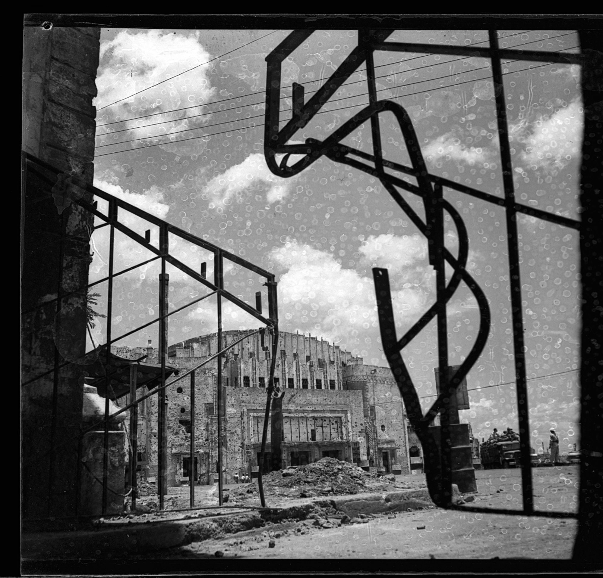 A black-and-white photograph of the Metropolitan Theatre, seen framed through the Insular Ice Plant Gate. To the right, the Gate is more twisting than to the left. There are white circular squiggles on the image that is presumably from film degradation.