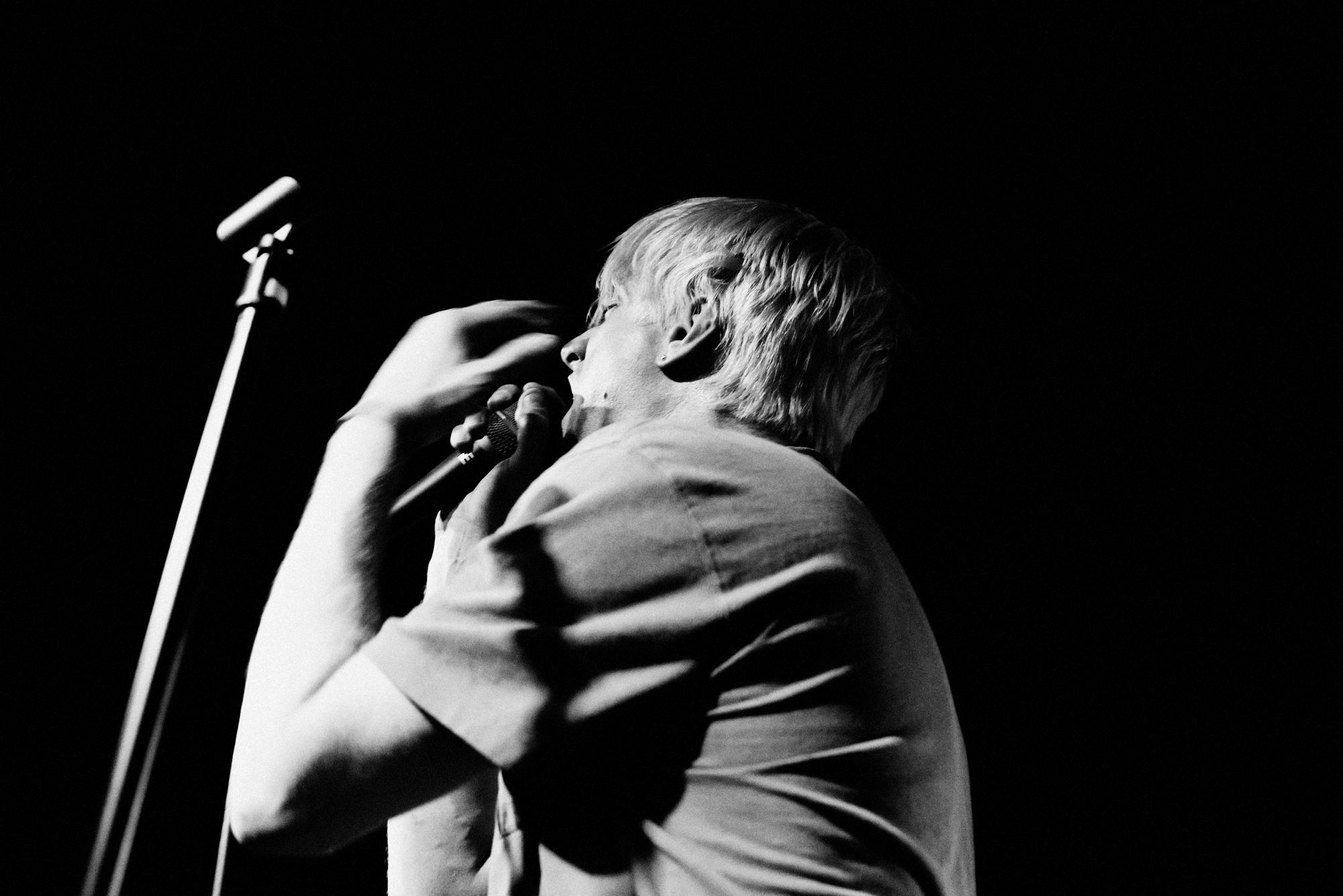 A black and white photo of a man singing into a microphone, mid-motion.