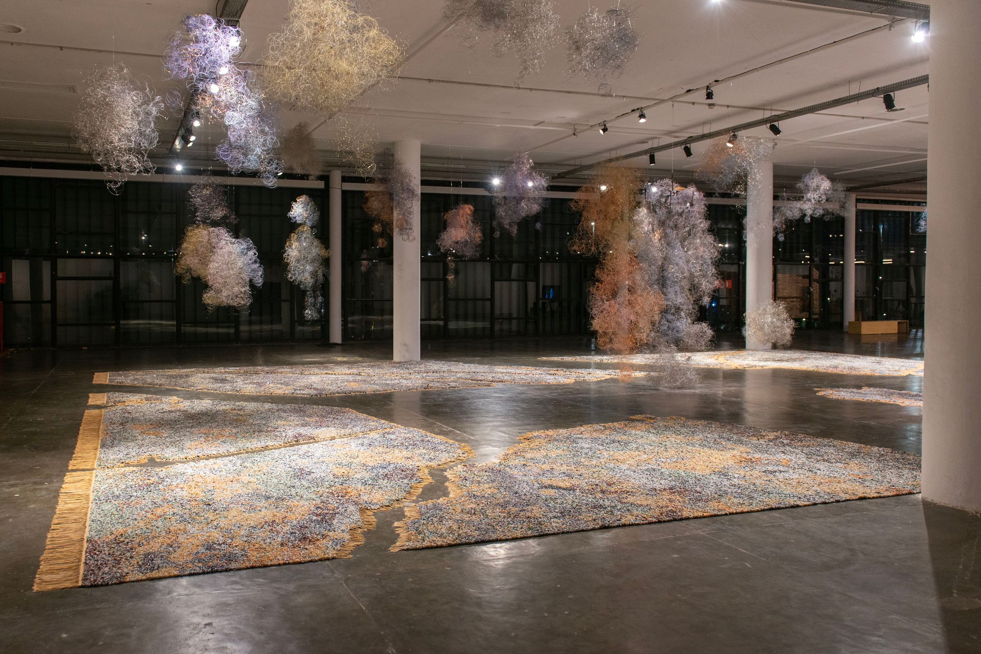 Pastel cloud-like structures extend from the ceiling of a gallery space, hanging above a yellow-toned carpet that covers the floor in irregular bursts.