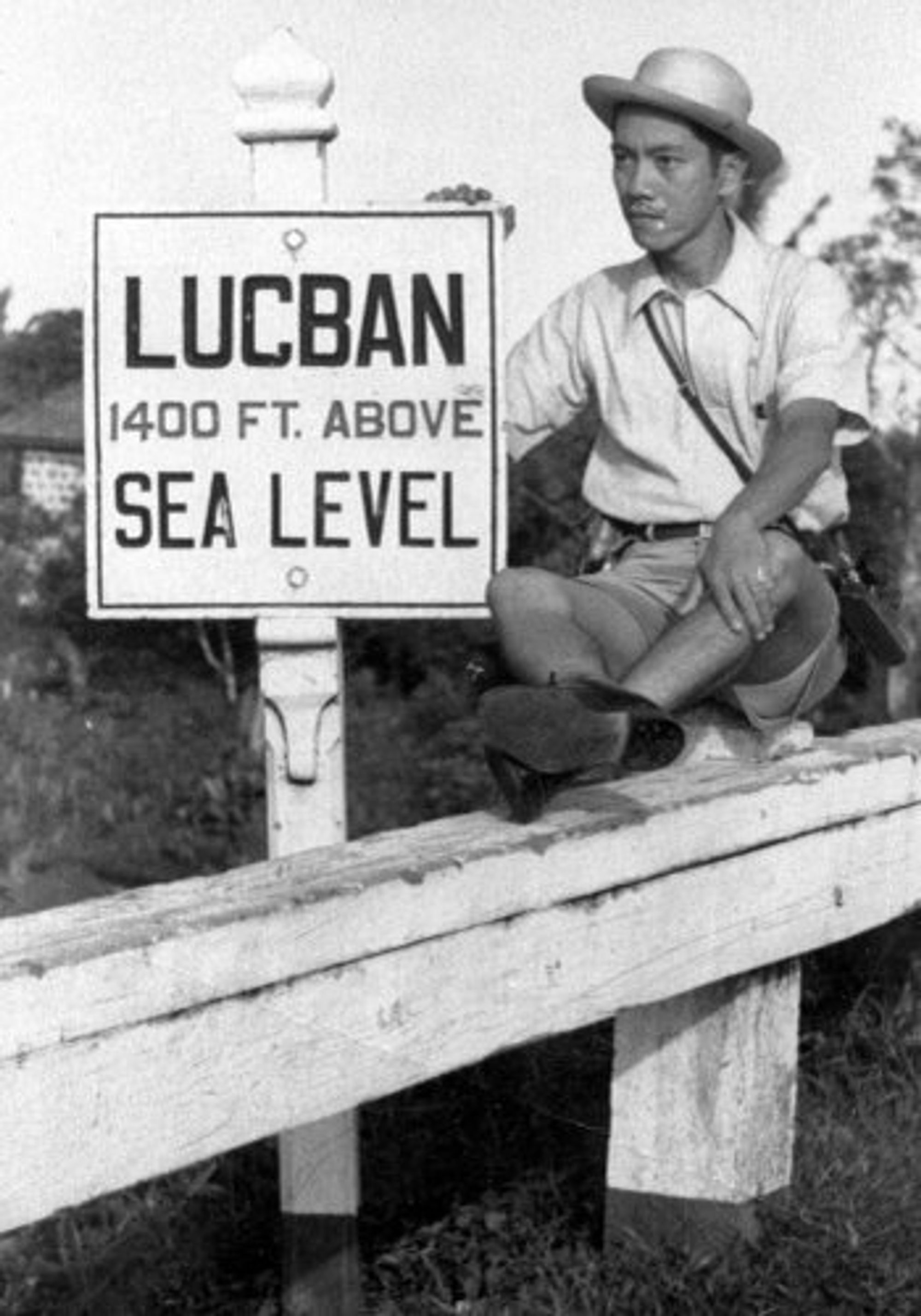 A photograph that depicts young Protomartir wearing a hat and a polo t-shirt. His expression is serious and he's looking off into the distance. His hand is resting on a sign that reads, "LUCBAN. 1400 FT. ABOVE SEA LEVEL"