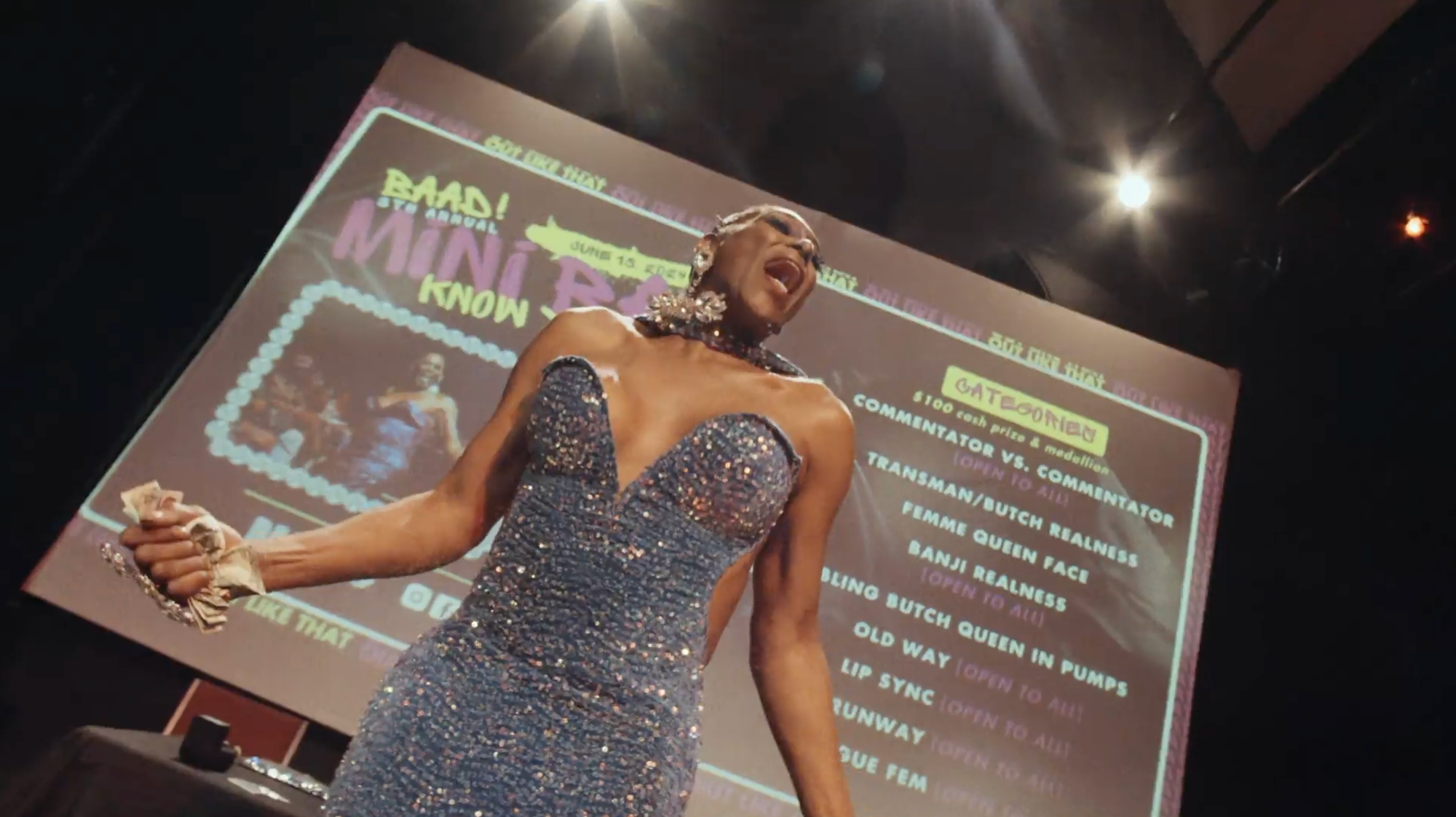 A drag queen singing at a ball show in a sequined blue gown with the stage screen behind her