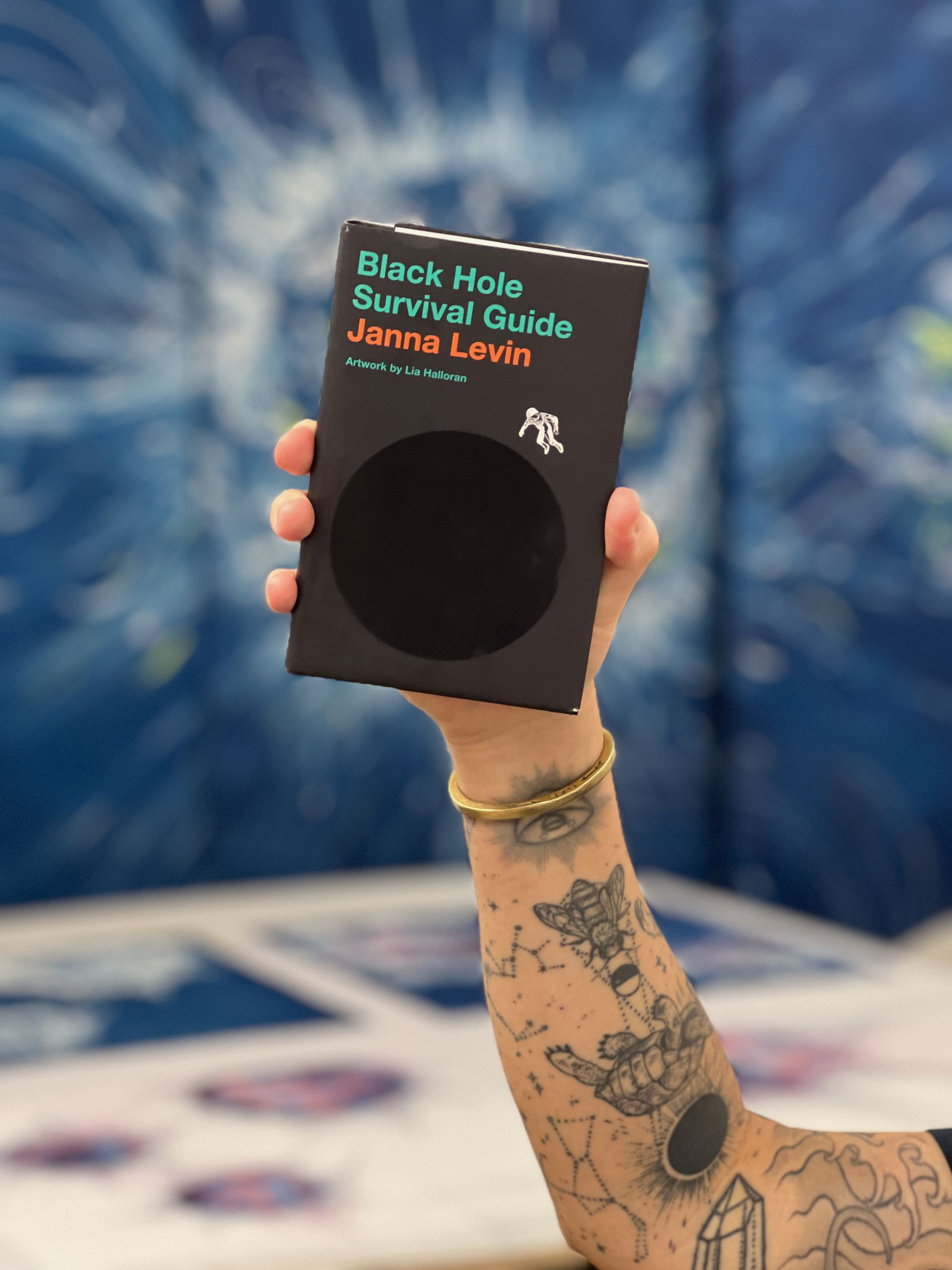 A tattooed arm holding up a copy of the book, Black Hole Survival Guide