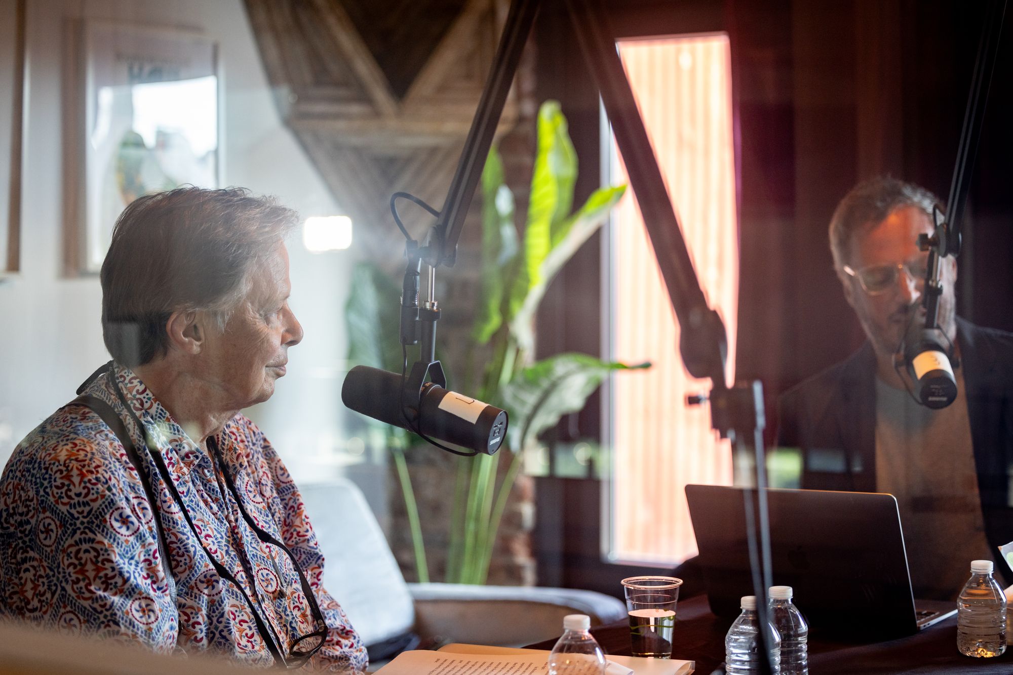 A photograph of two men, taken through the glass of a radio booth, speaking to each other into microphones. 