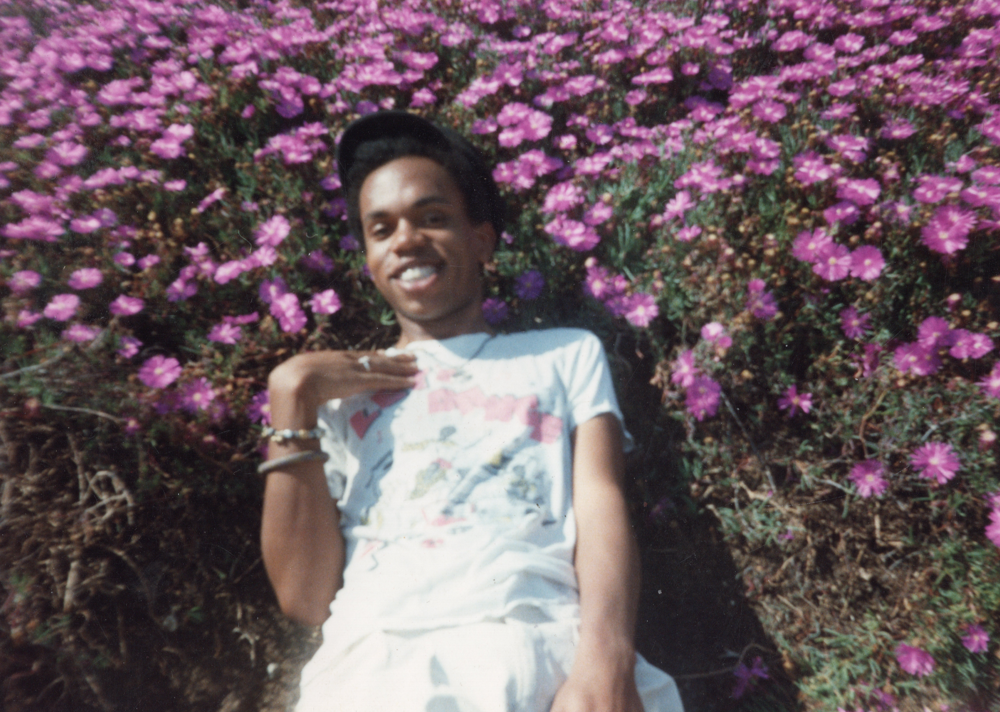 A young Sean DeLear wearing a cap, lying down in a bed of purple flowers with one hand across his chest. 