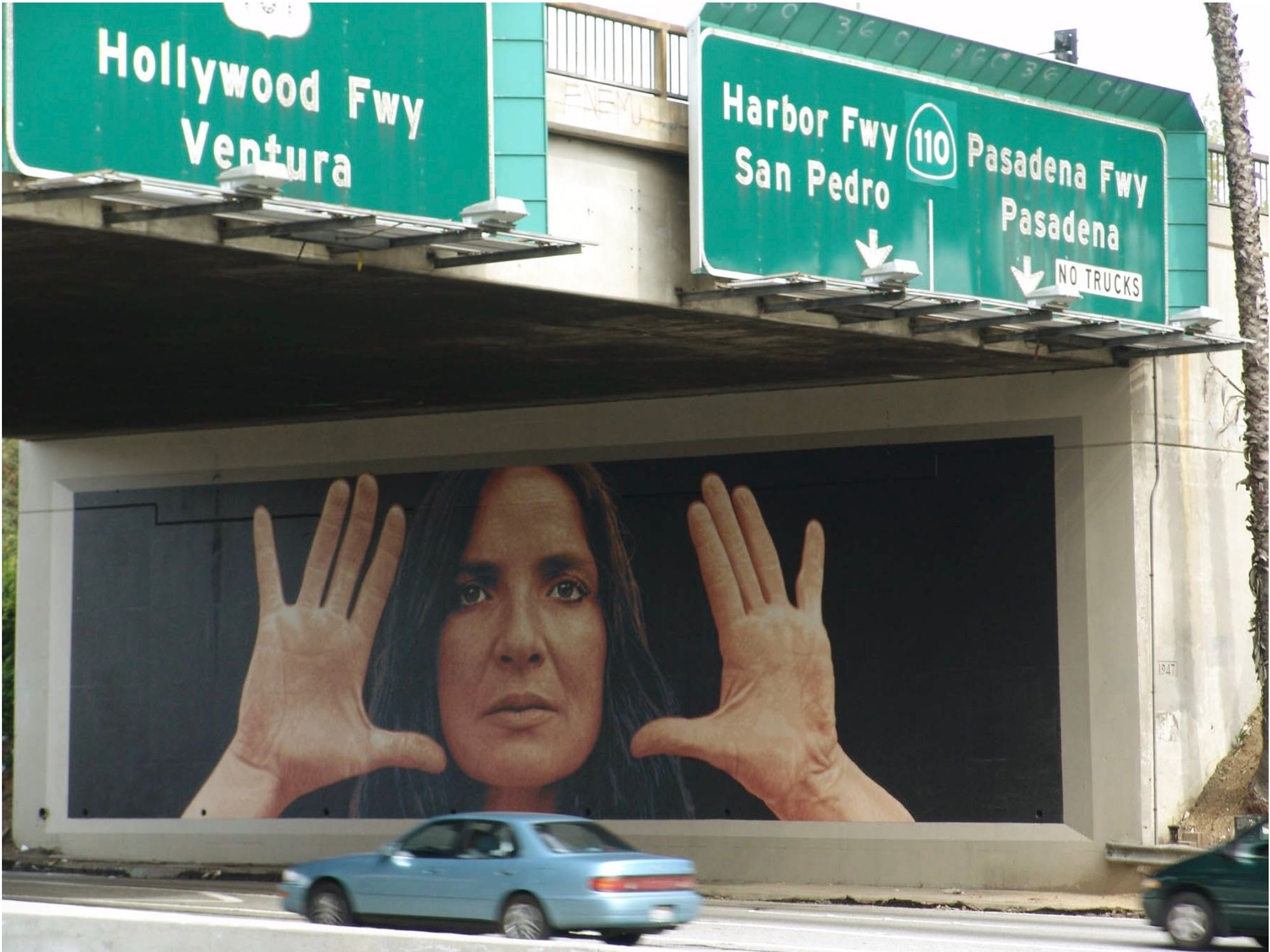 A mural on the side of the freeway depicting a woman staring out from between her hands.
