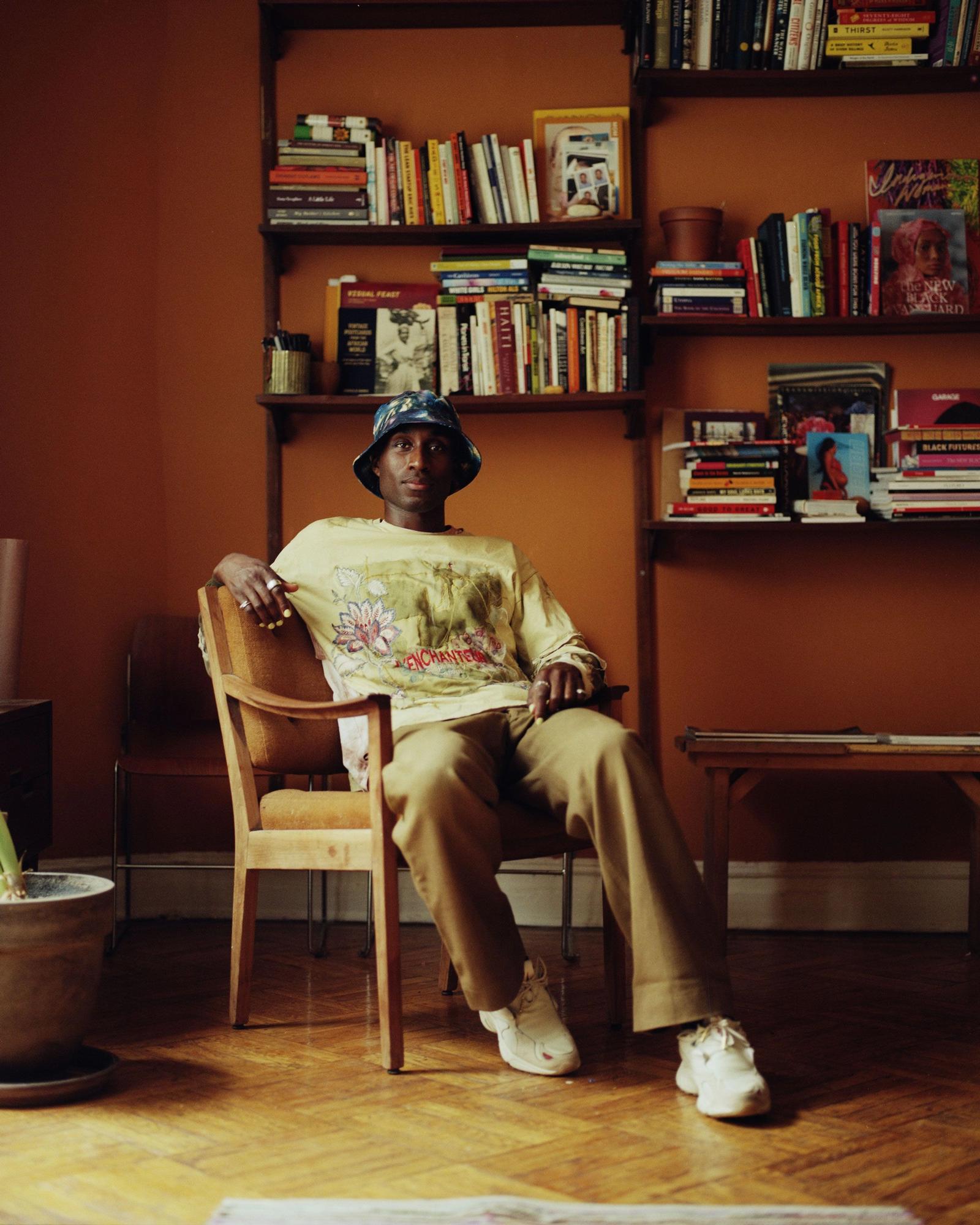 Devonn Francis sitting in front of bookshelves, wearing tan clothing and a blue bucket hat.