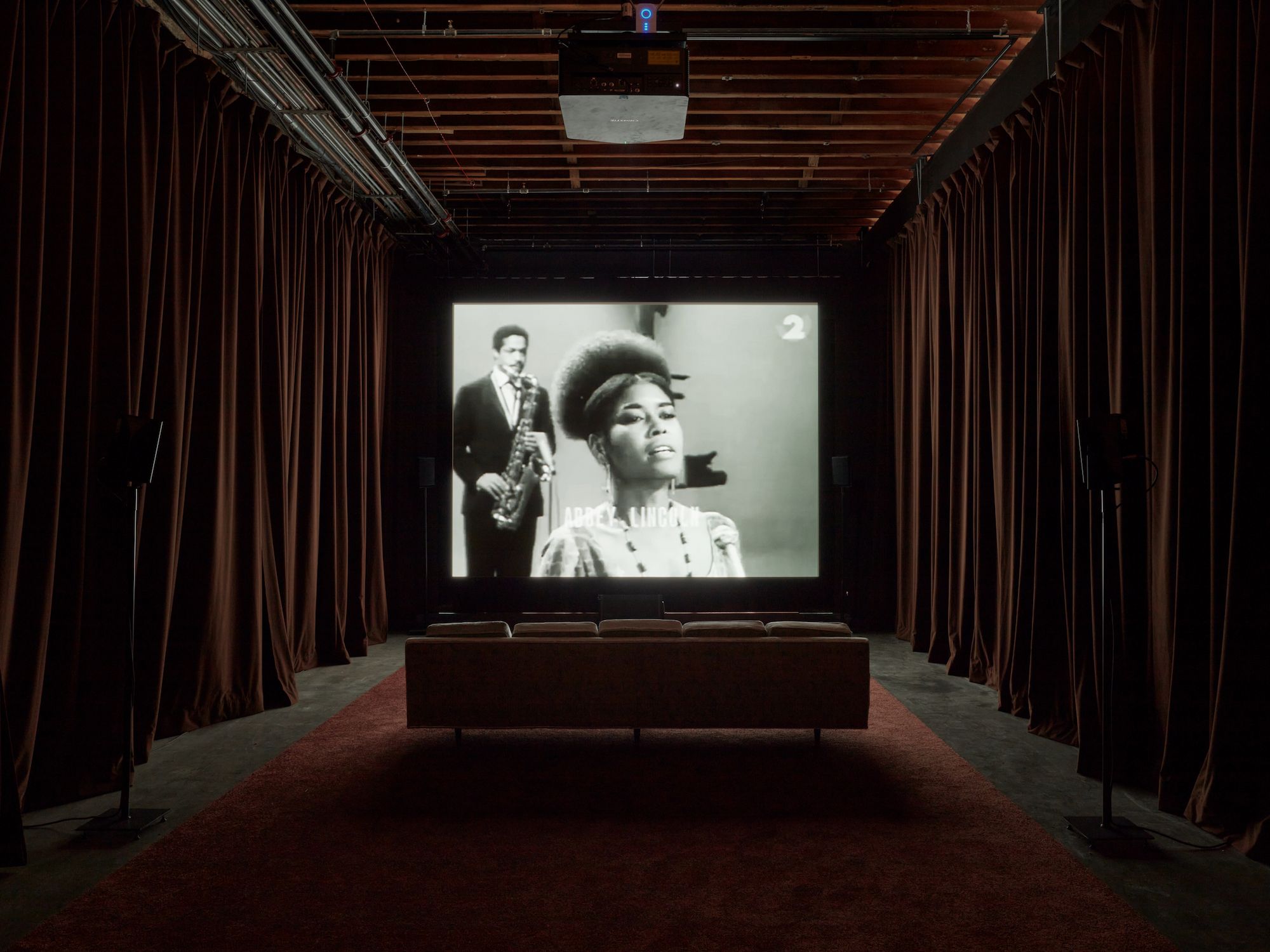 An installation view of inside of a black box makeshift theatre where a screen in the center displays a black-and-white video of a black woman singing while a black man plays saxophone in the background