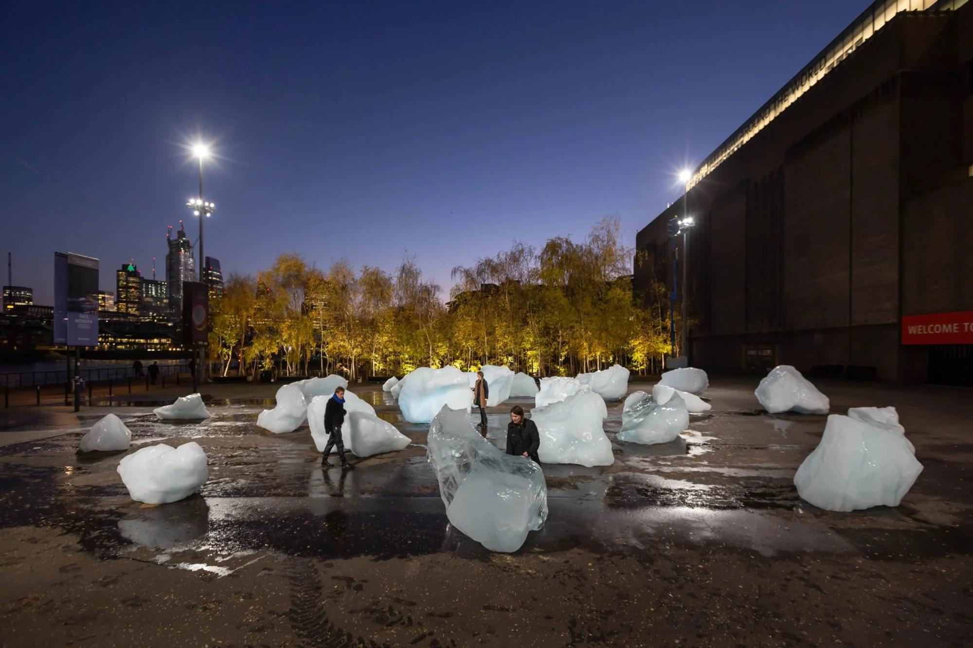 A photograph of people walking around melting glaciers that have been dropped into an urban landscape. 