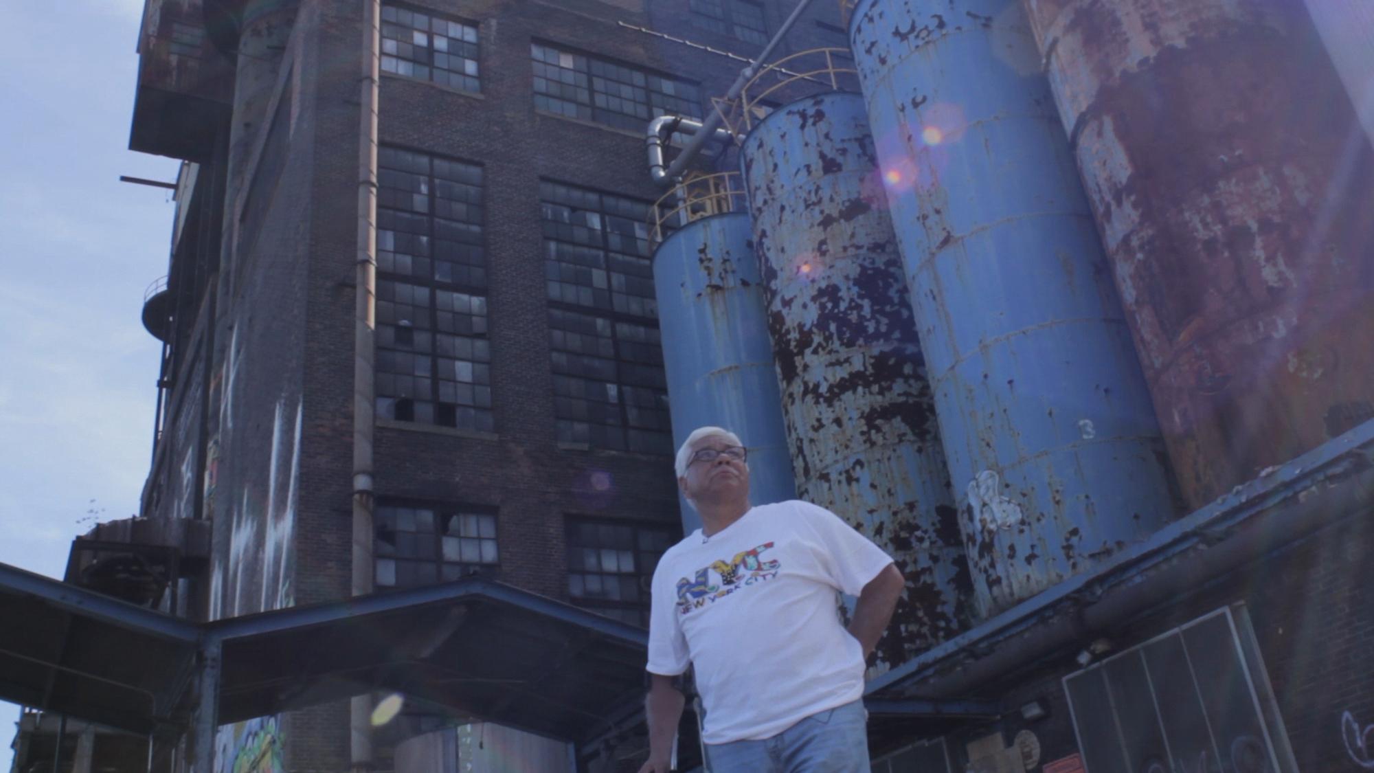 An older man (Frank Ortiz) wears a white shirt with the letters NYC on them and blue jeans stands in front of an industrial building, the Domino Sugar refinery.
