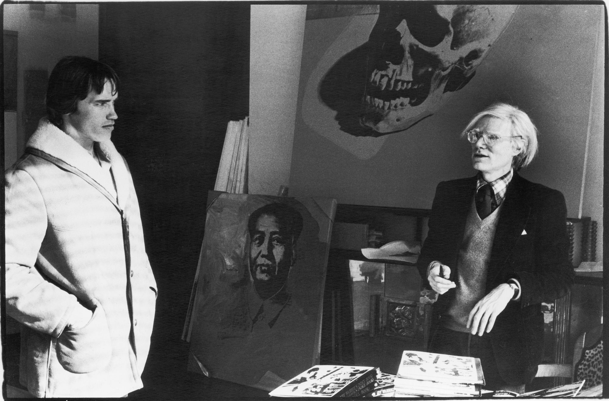 A young Arnold Schwarzenegger stands in an art studio, looking intently at Andy Warhol.