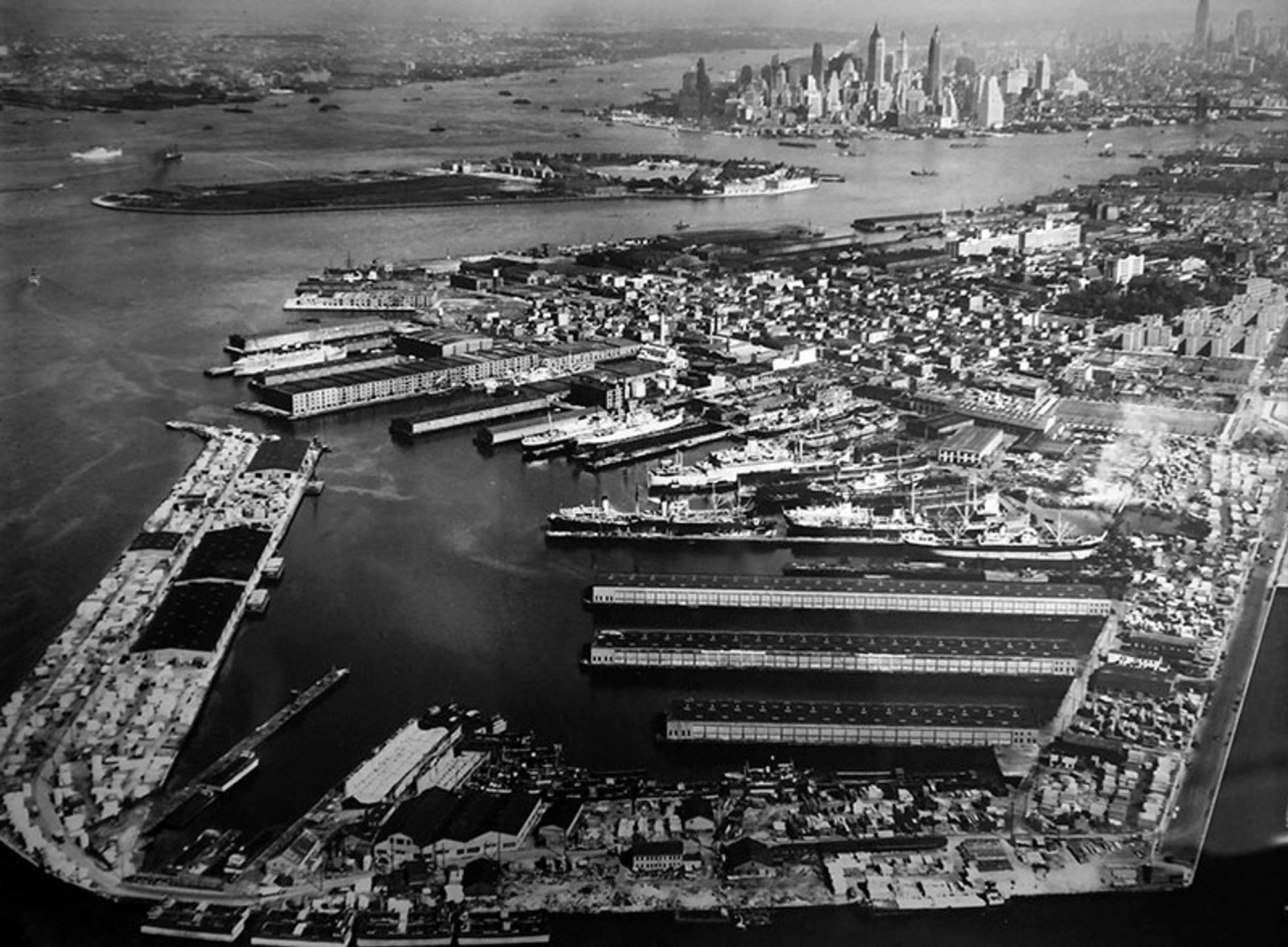 A black-and-white aerial view of Red Hook, with the working waterfront and Erie Basin Terminal in the foreground, NYCHA public housing projects to the right, and Lower Manhattan and Governors Island seen in the distance. 