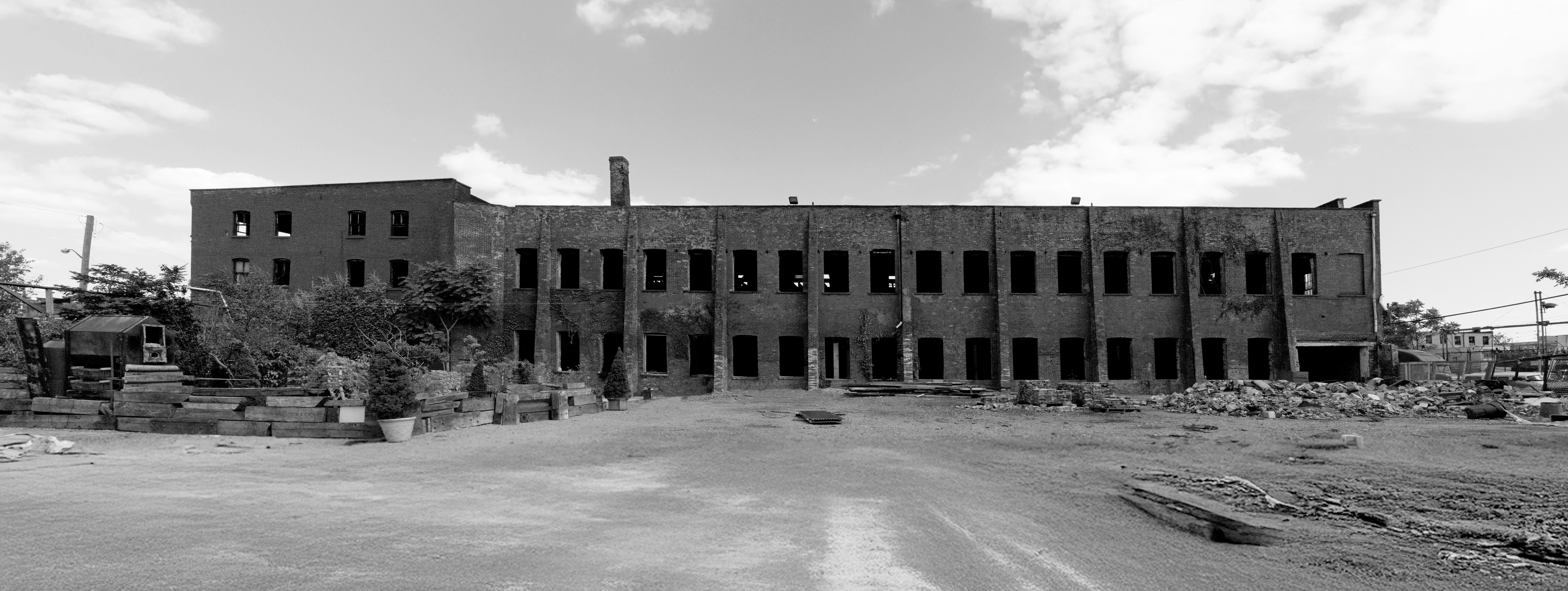 A black-and-white photograph of a hollowed-out industrial building, made of brick and wider than it is tall, set upon a concrete ground and photographed against a gray sky. 