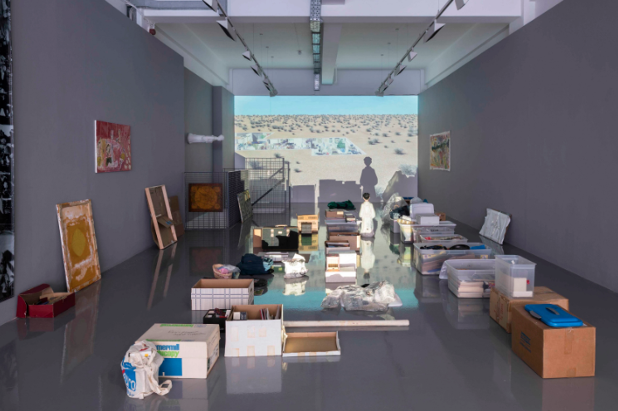 In a gallery space, a collection of objects are arranged as if stored in some sort of garage—artworks leaning against a wall and papers stacked on top of one another in boxes. A desert landscape is projected against the back wall of the gallery space. 