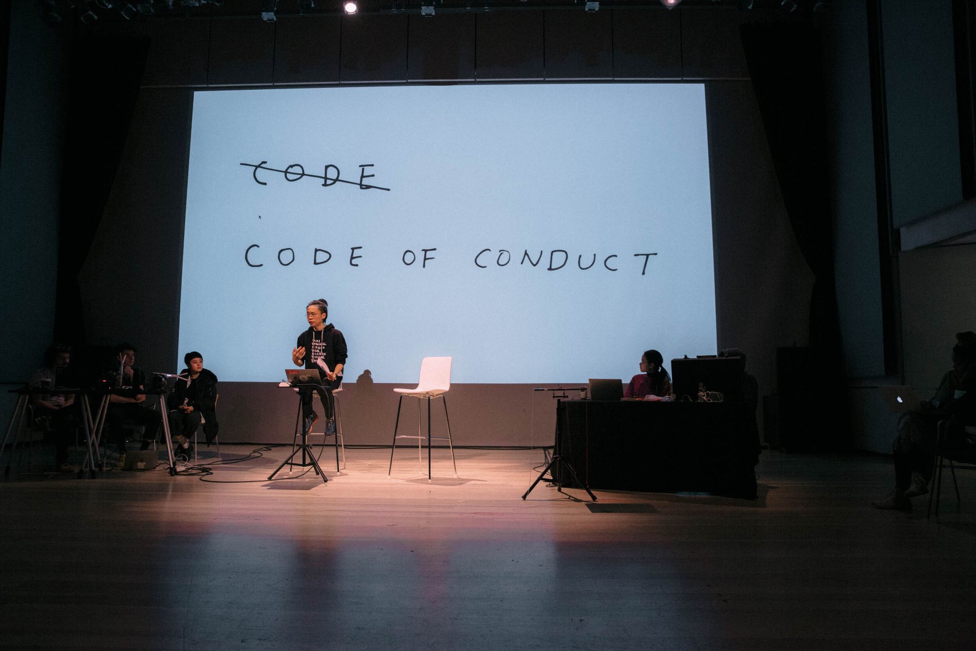 Taeyoon is spotlit and wearing a black sweatshirt, sitting on a chair that's on a stage. Behind him, a large screen shows a handwritten images where the word "code" is crossed out, and "code of conduct" is written below.