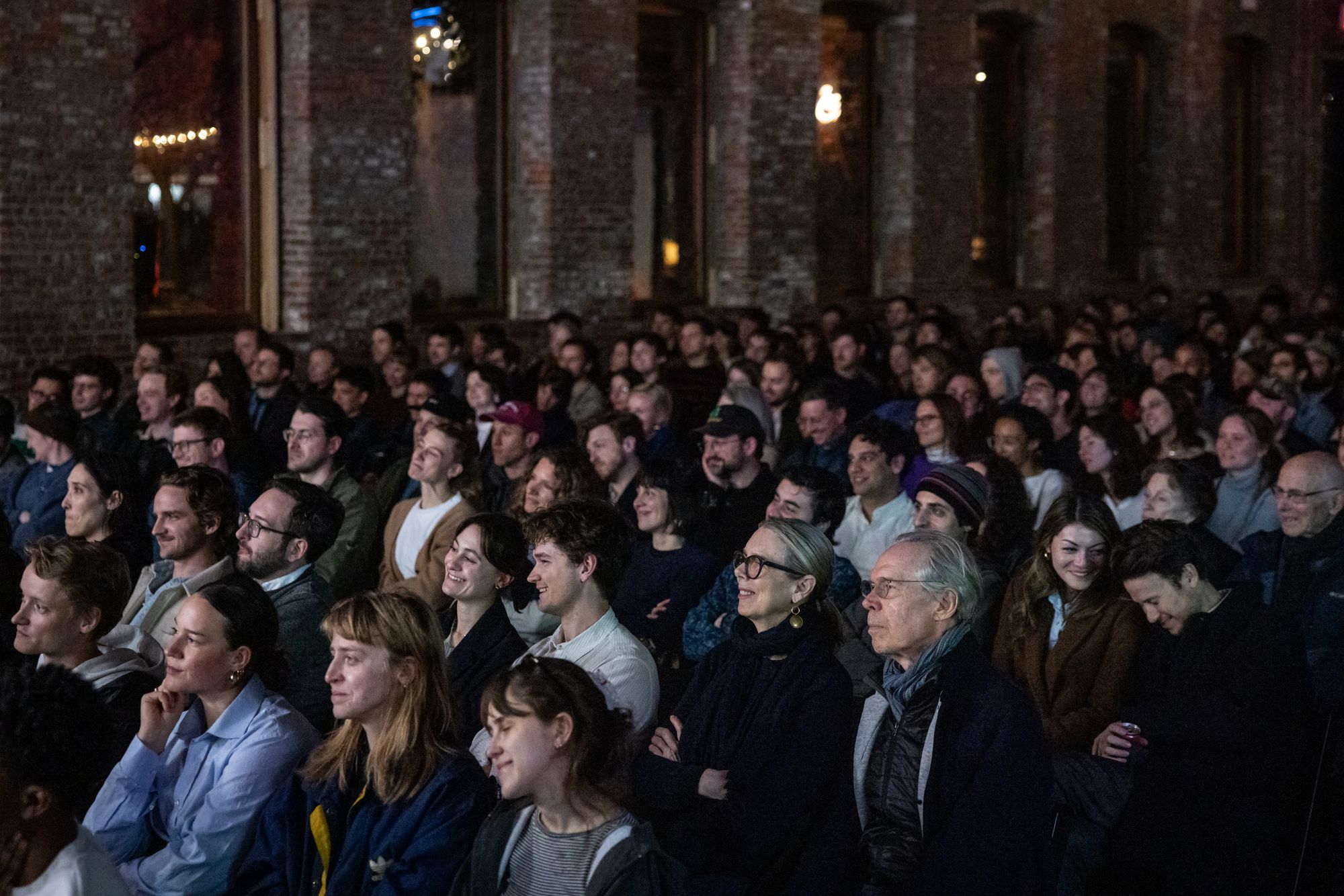 An audience in a dark room, all looking at a subject beyond the frame, some of them smiling and laughing.