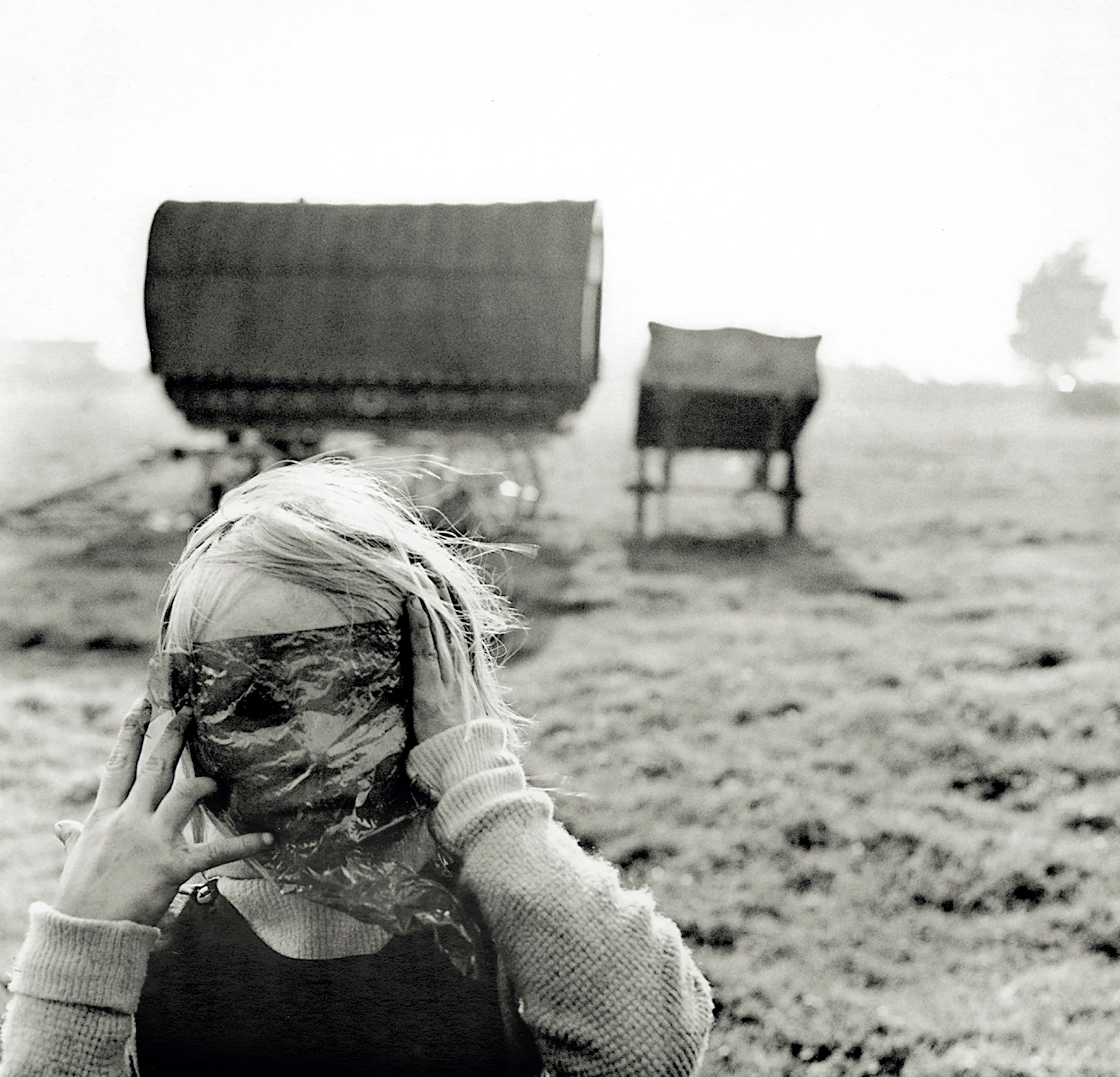 A black and white photograph of a blond child covering her eyes with a translucent piece of plastic, with the wind blowing in her hair. She stands in front of a covered wagon in an open field. 