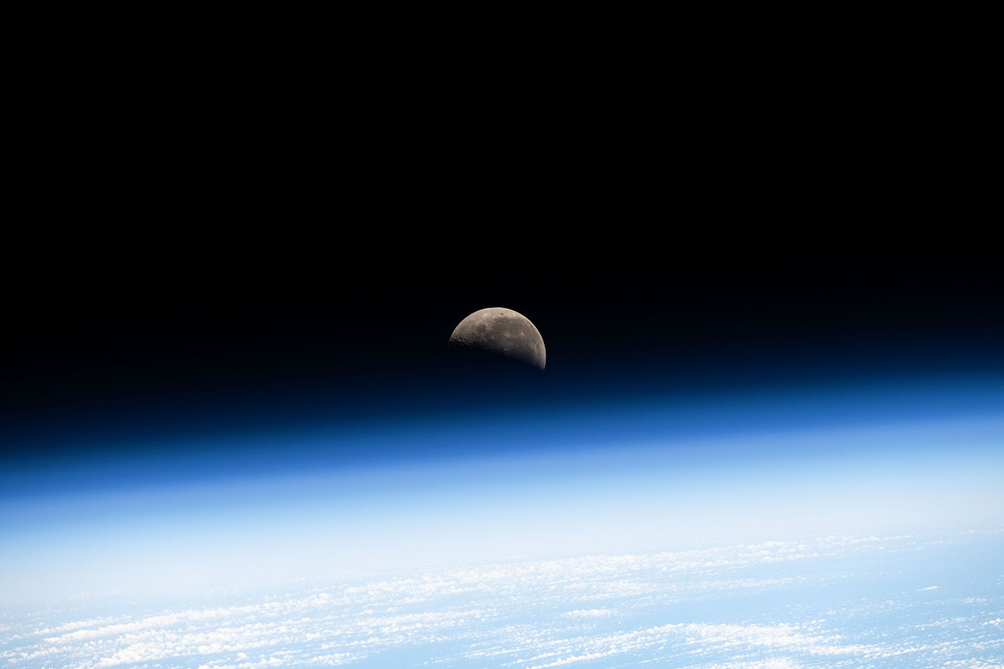 The Earth's atmosphere creates a blue, shiny bottom-half on the picture. Half of the moon is seen amidst the blue haze, right at the center, beneath the rest of dark space.