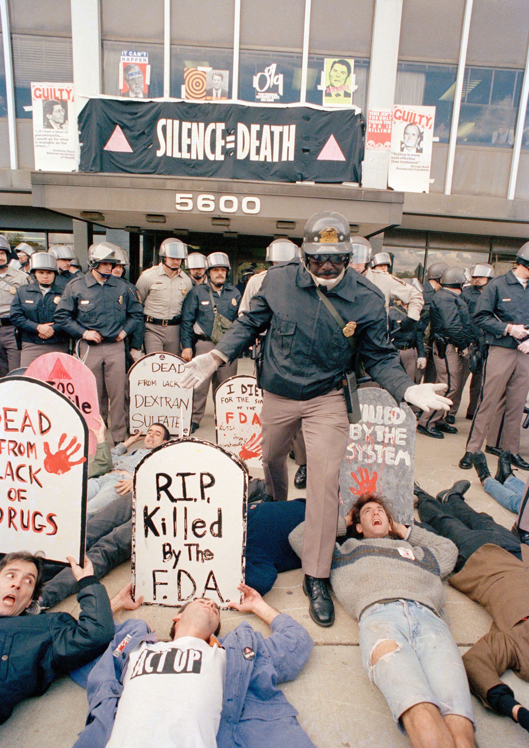 A police officer poses above several people laying on the ground with tombstones they've drawn protesting the FDA.