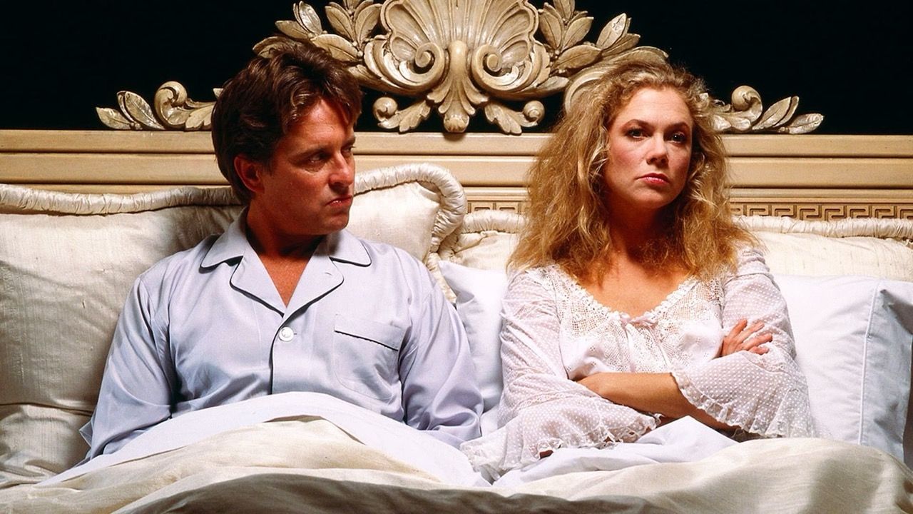 In a film still from War of Roses, a married couple sits upright in bed, visibly angry at one another.