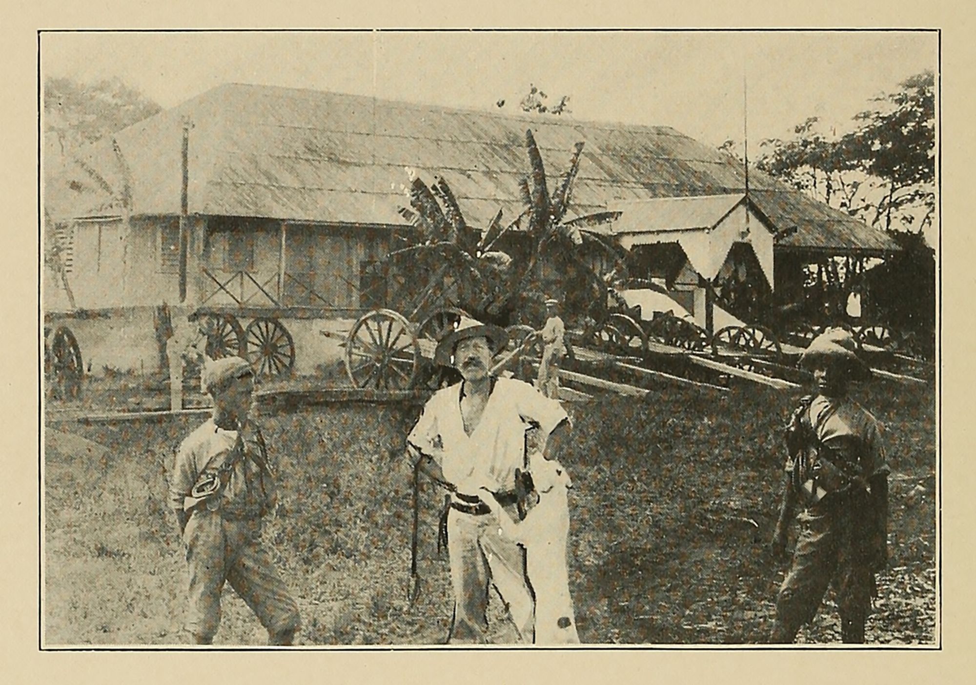A white man in a hat stands in a tropical setting, flanked by two younger men with darker skin.