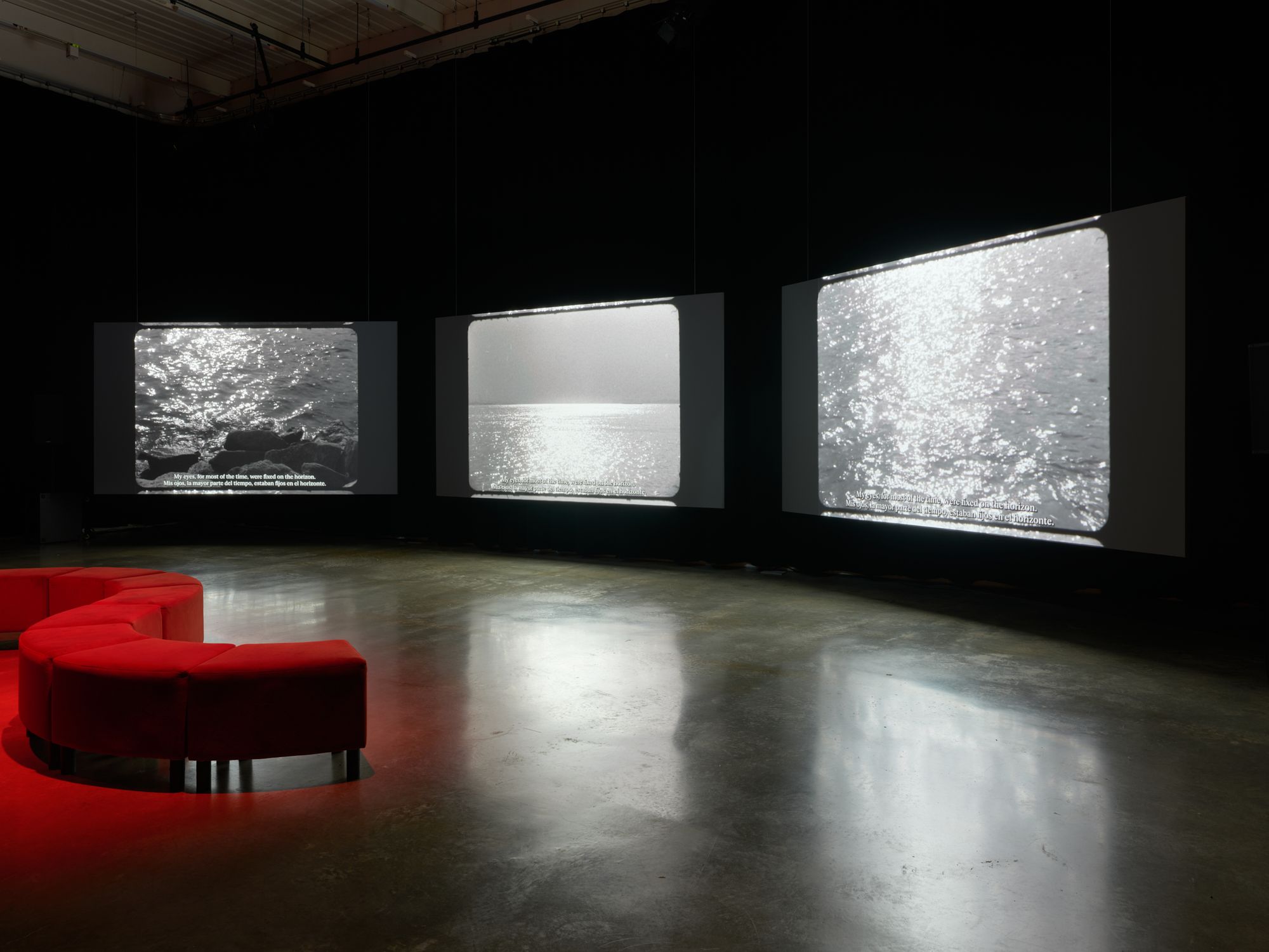 A red couch sits in front of a triptych of screens, each projecting some image of glittering oceans. 