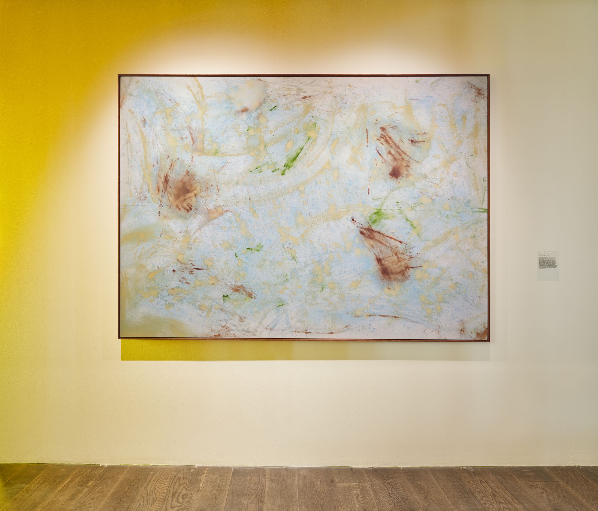 A painting featuring smudges of light blue, green, and brown pigment hangs on a wall lit in swatches of yellow light