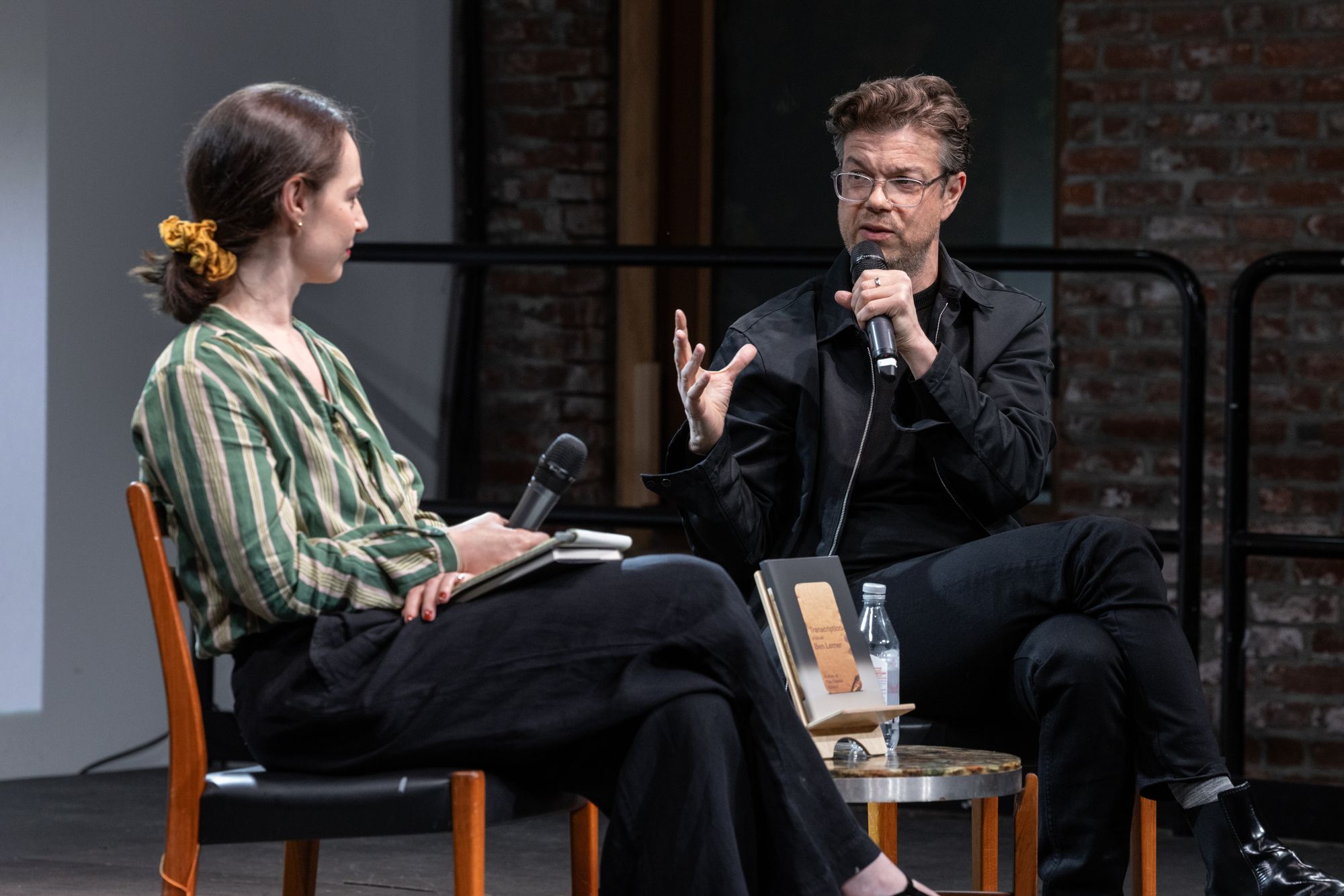 Alexandra Schwartz and Ben Lerner sit on stage, speaking to each other into microphones, with a backdrop of the Transcription book cover looming behind them.
