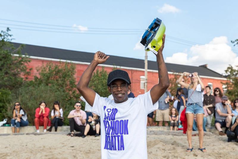 A boy wearing a white shirt that reads "Red Hook Regatta" in blue text holds a boat over his head victoriously, as folks in the background sitting on the beach cheer him on 