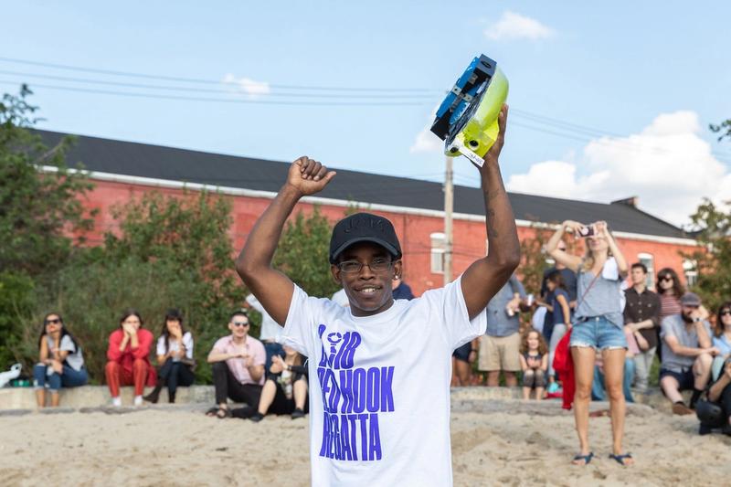 A boy wearing a white shirt that reads "Red Hook Regatta" in blue text holds a boat over his head victoriously, as folks in the background sitting on the beach cheer him on