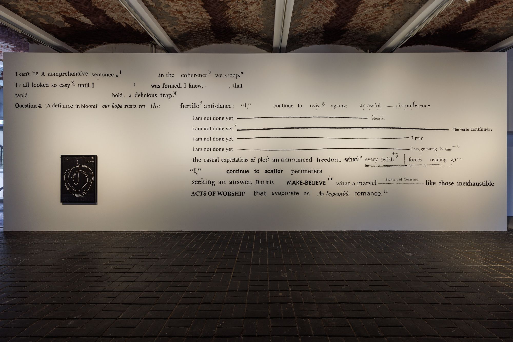 An installation view of a white wall covered in black text, with lines like "I can't be A comprehensive sentence."