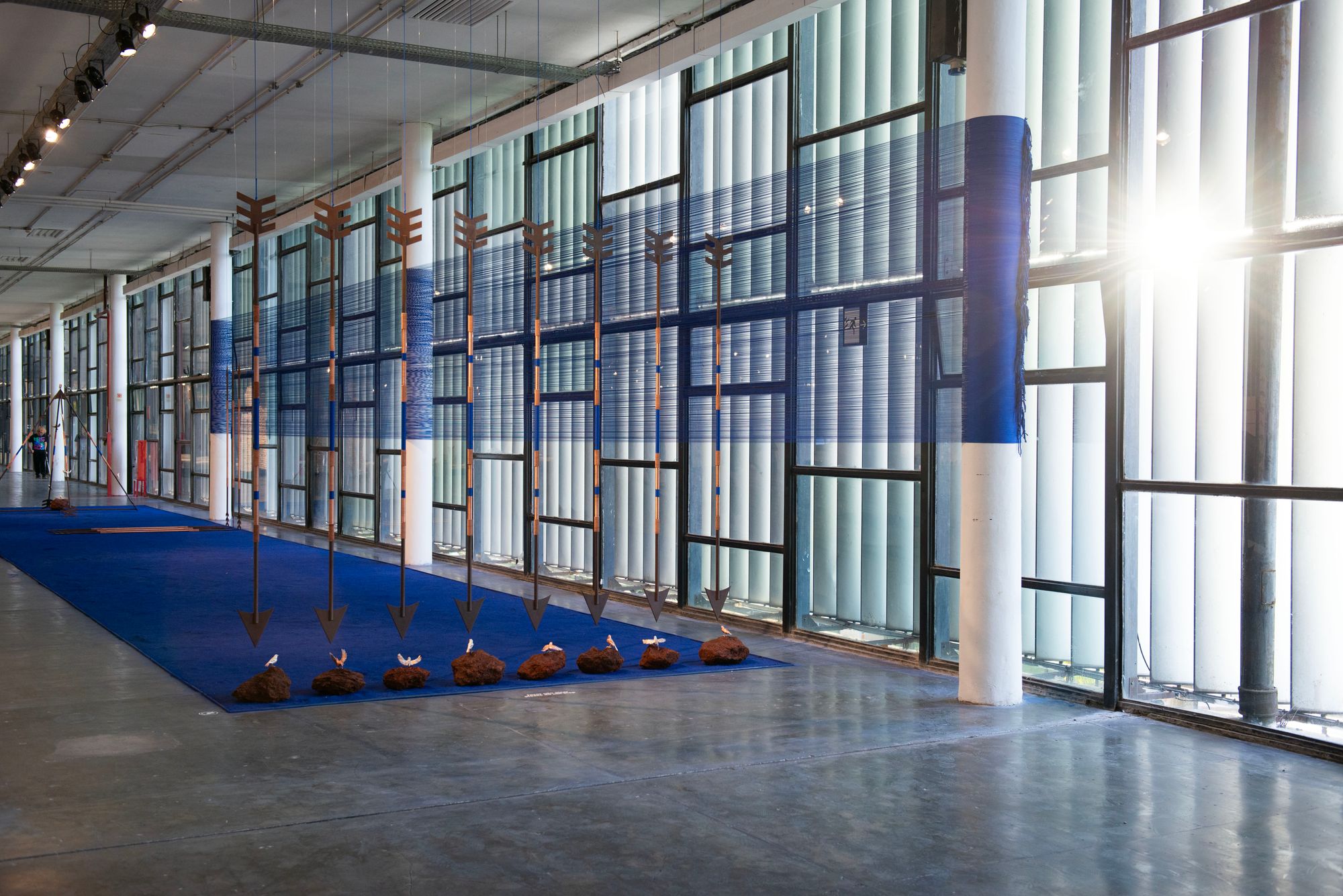 A sculpture situated on a concrete floor that looks upon a floor to ceiling window, where eight parallel arrows point downwards, towards small doves resting upon irregular rocks, which sit at the very end of a long blue rug.