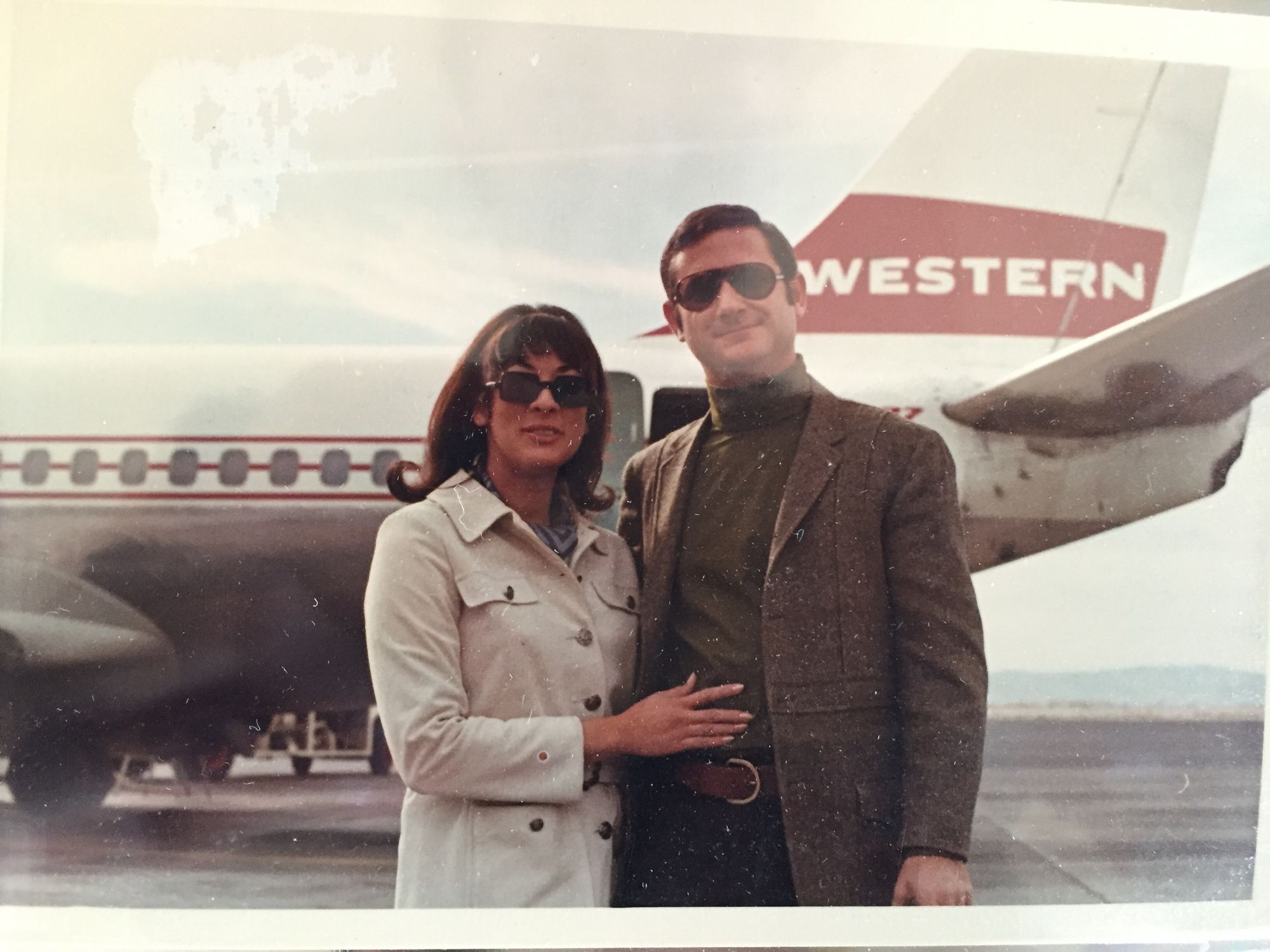 A man and a woman standing outside of a plane wearing outfits reminiscent of the '60s and '70s.