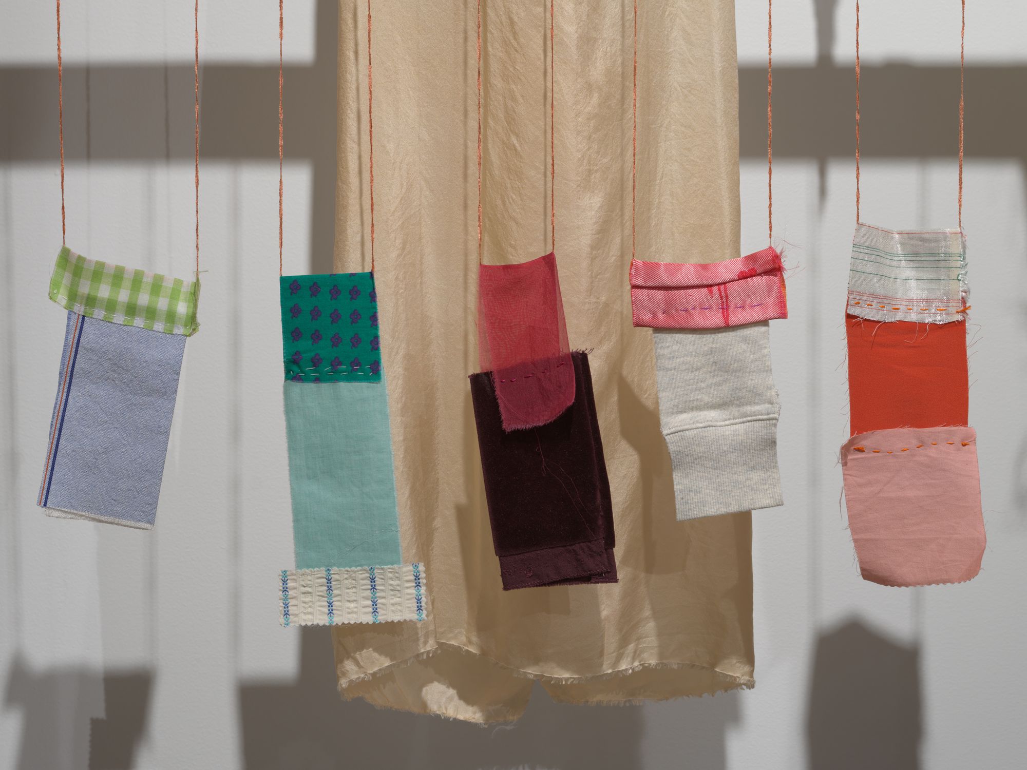 Delicate pouches made from silky material dangling from a wooden plank in a gallery space.