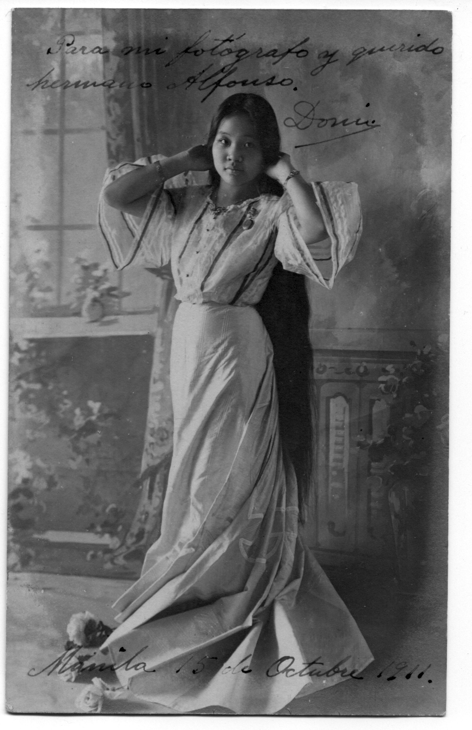  A portraiture of a woman who is wearing a flowing dress and posing for the camera. Both of her arms are raised as she coyly tucks her hair back. She looks directly at the camera. There is cursive script above and below the portraiture.