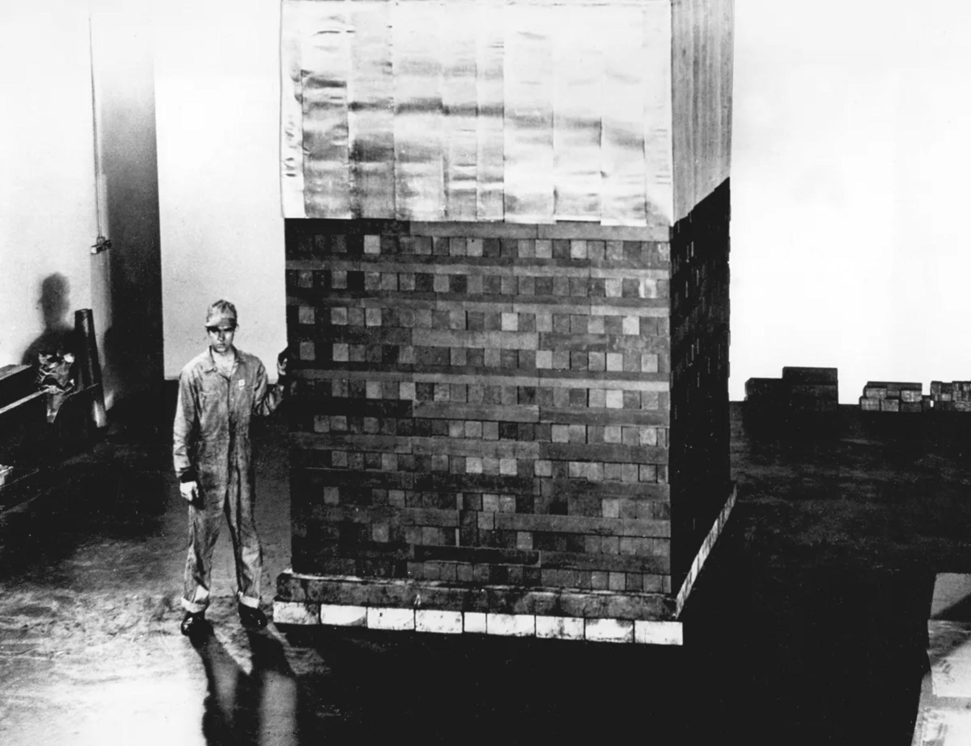 A black and white image of a man sitting next to what looks like a uniformly spaced pile of bricks. 
