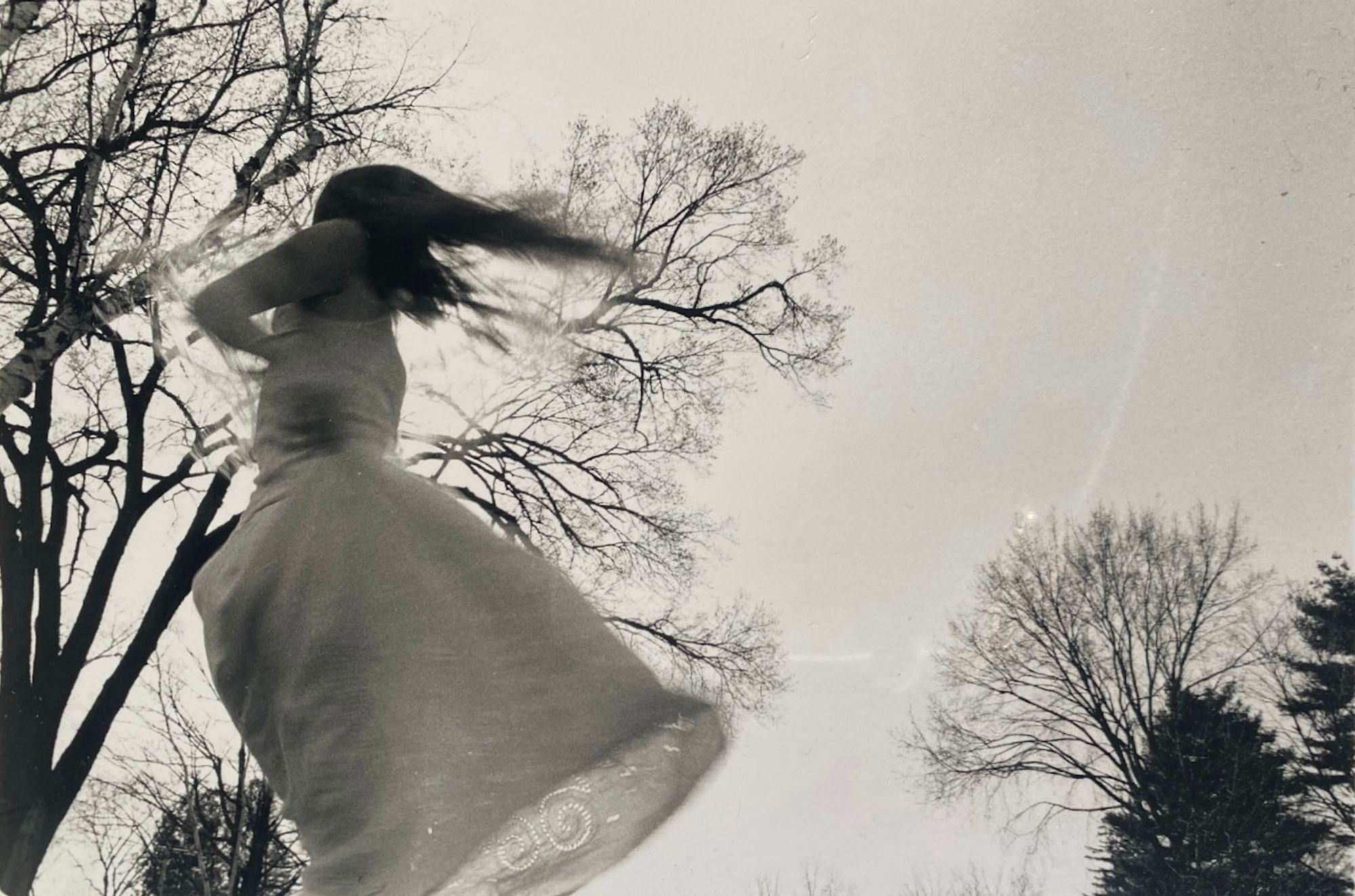 A spinning figure in a white skirt swirls against the silhouette of trees against a grey sky.