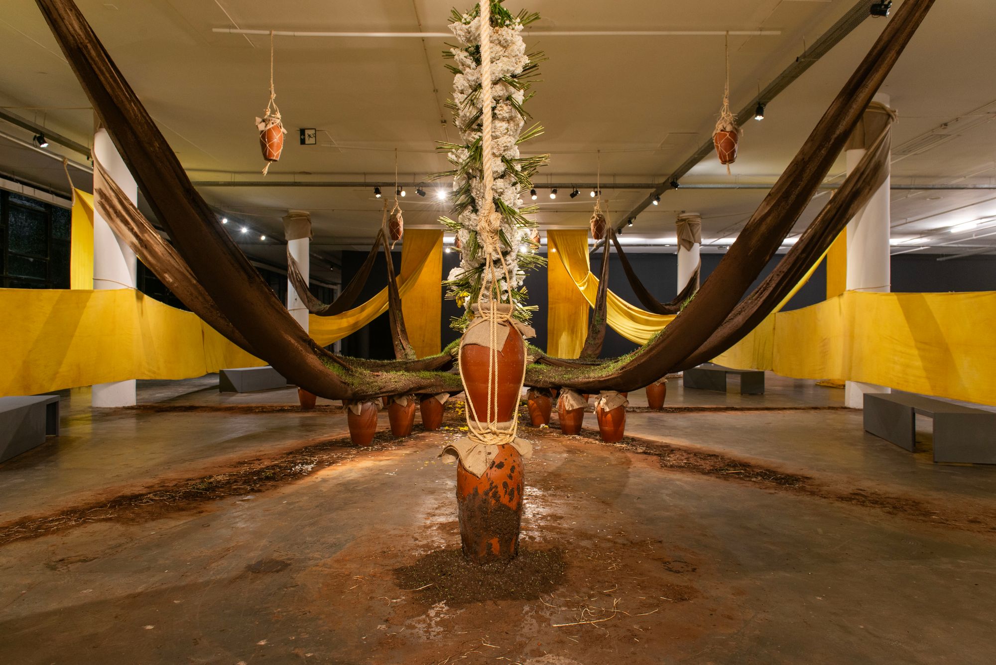 A clay pot rests at the center of a vibrant still life, wherein brown and yellow cloths are festooned from the ceiling but convene inwards towards the center.