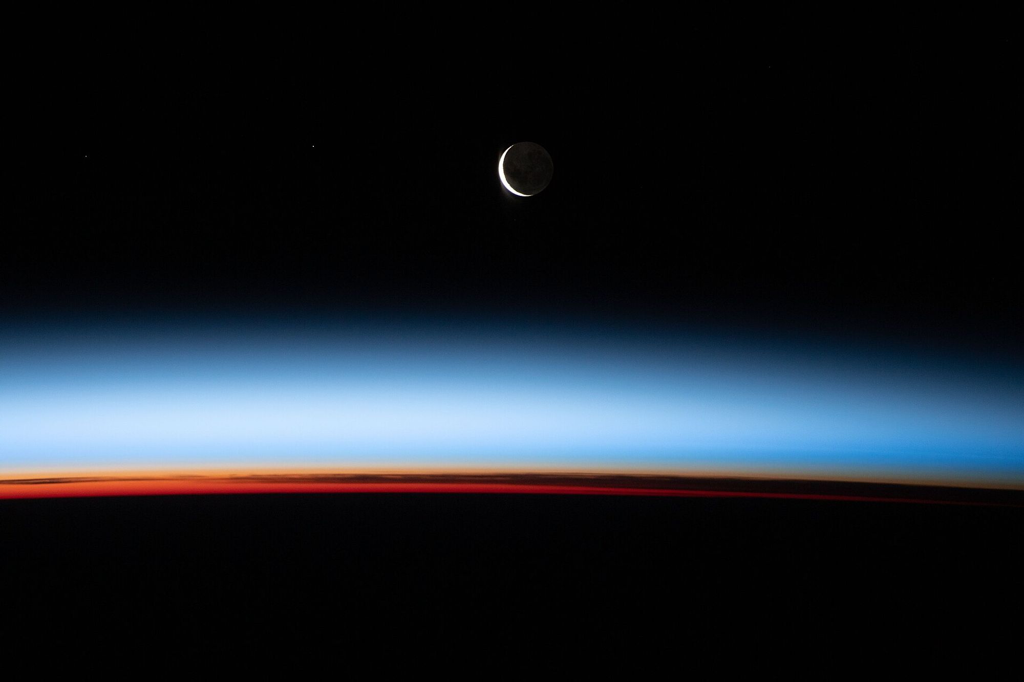 A blue, hazy band cuts the image in the middle. At the top, a sliver of the moon is seen against a dark space background. At the bottom, a slim band of orange, or sunrise on Earth, cuts across the image.