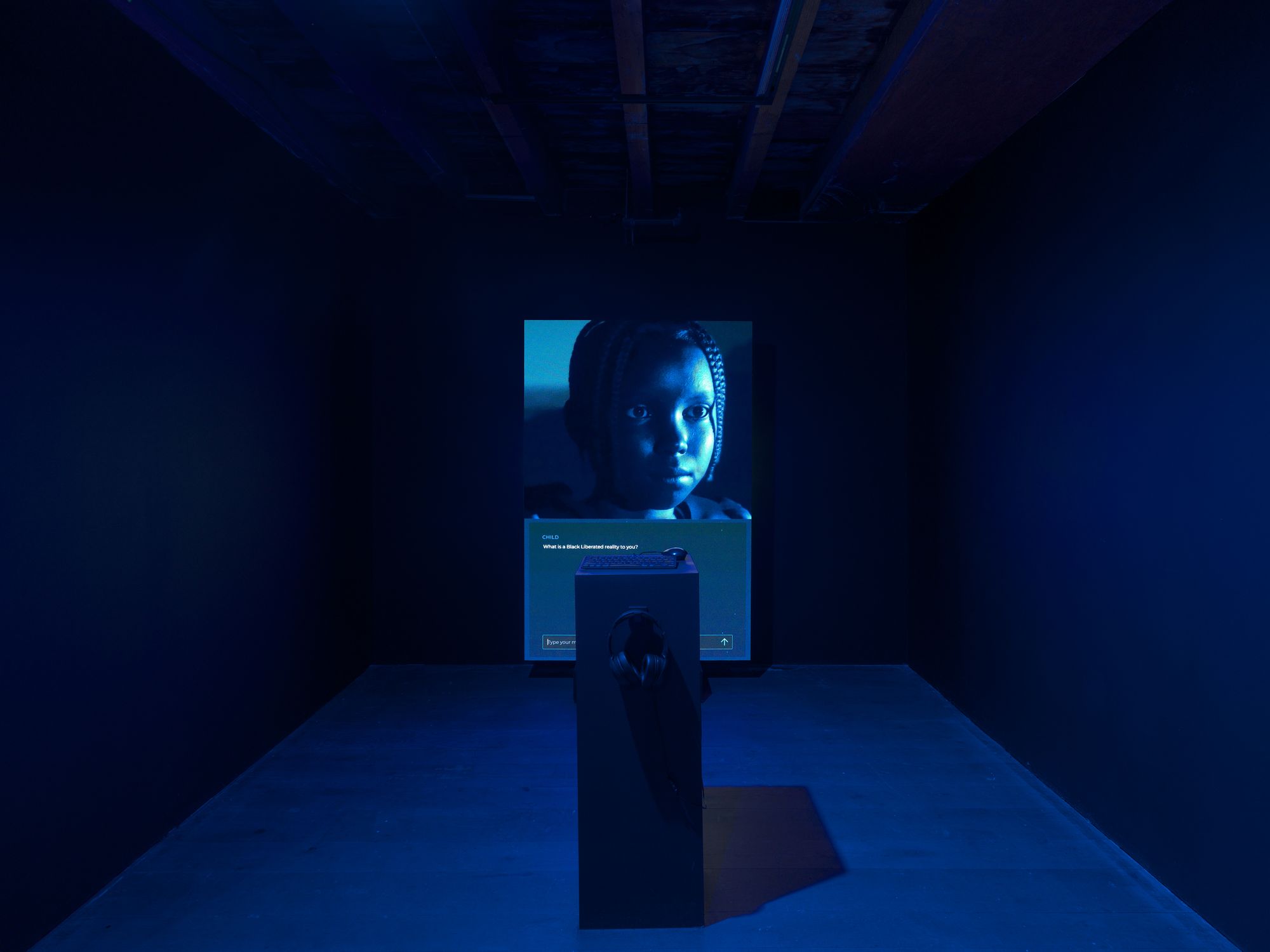 A screen in a dark room that features an image of a young black girl with a query written below that reads: "What is a Black Liberated reality to you?"