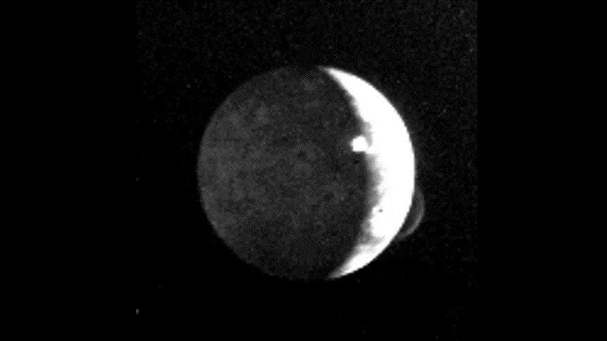 A grainy black and white photos shows Io pockmarked by a bright white plume of volcanic emission.
