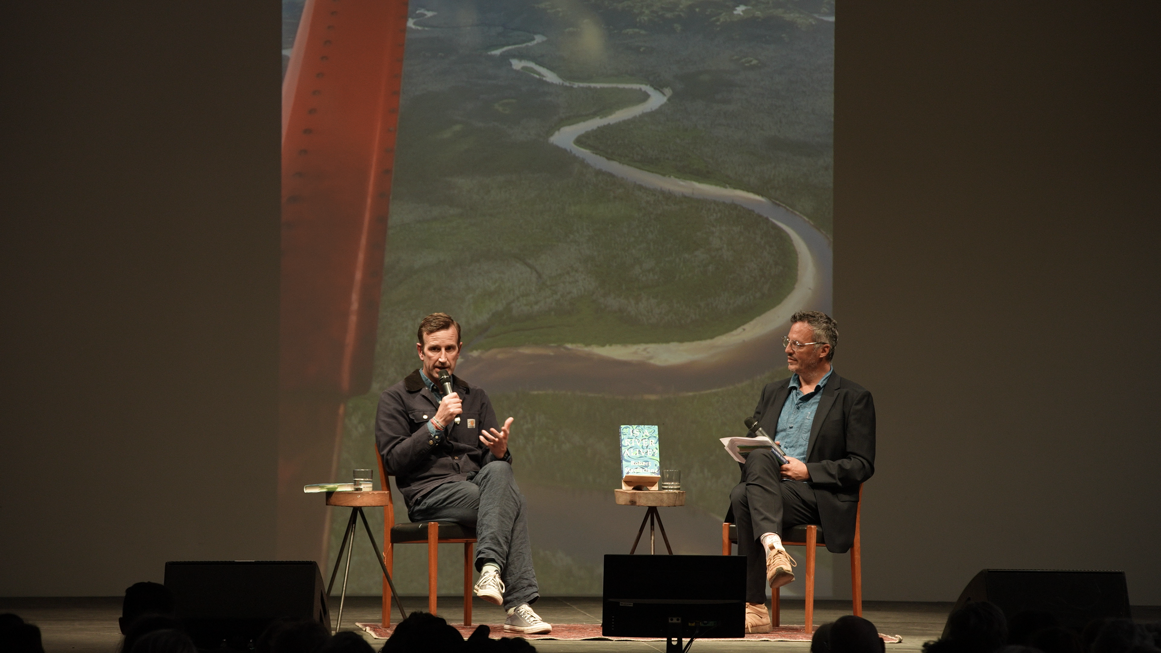 Two men sit on stage and speak into microphones, with a picture of a river projected behind them. 