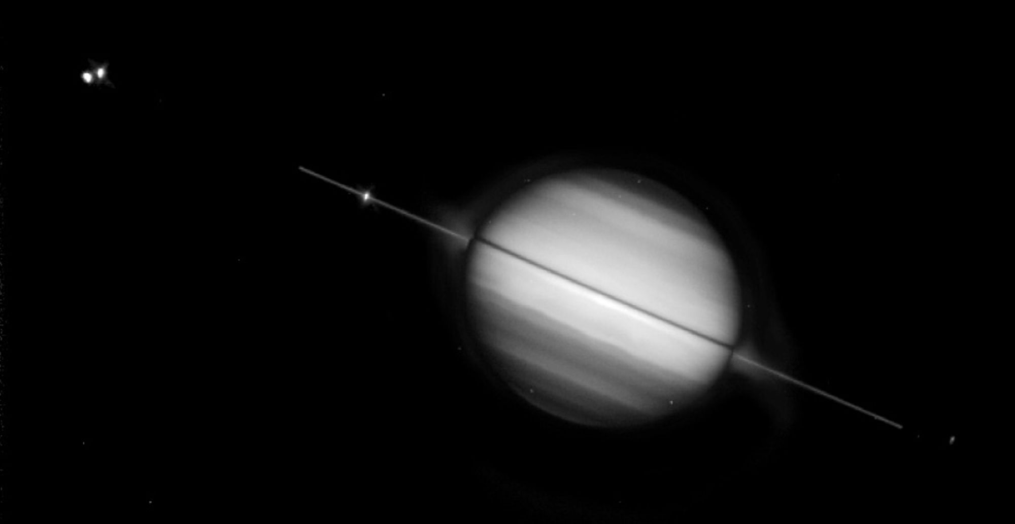 A black-and-white photograph of Saturn, with its rings seen edge-on, appearing as a thin line through the planet's middle.  