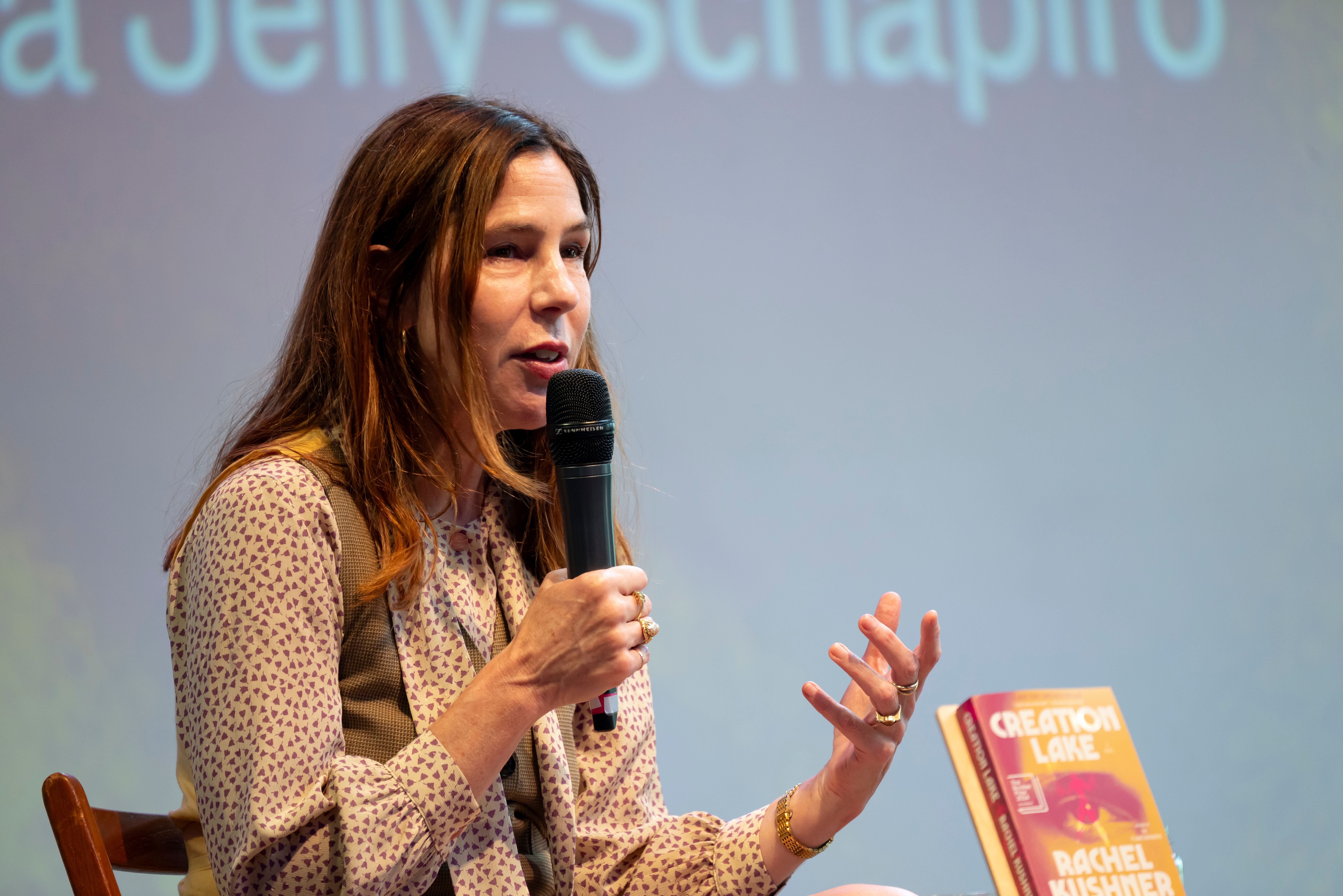A woman with shoulder-length brown hair speaks into a microphone, with the book Creation Lake held up on a small easel next to her.