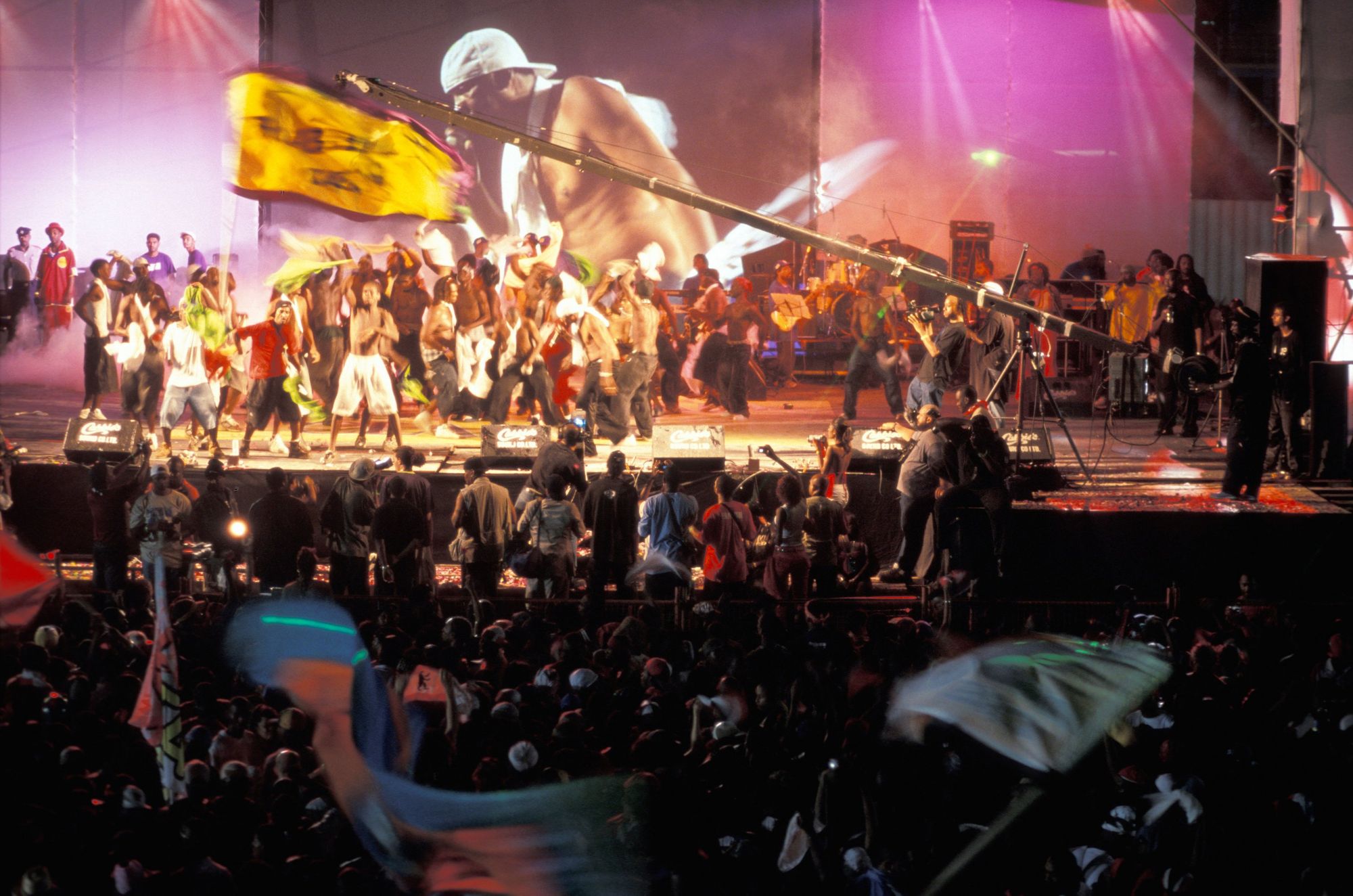 A photograph of a stage full of people, performing in front of a packed audience, with flags being waved in the air above the crowd. 