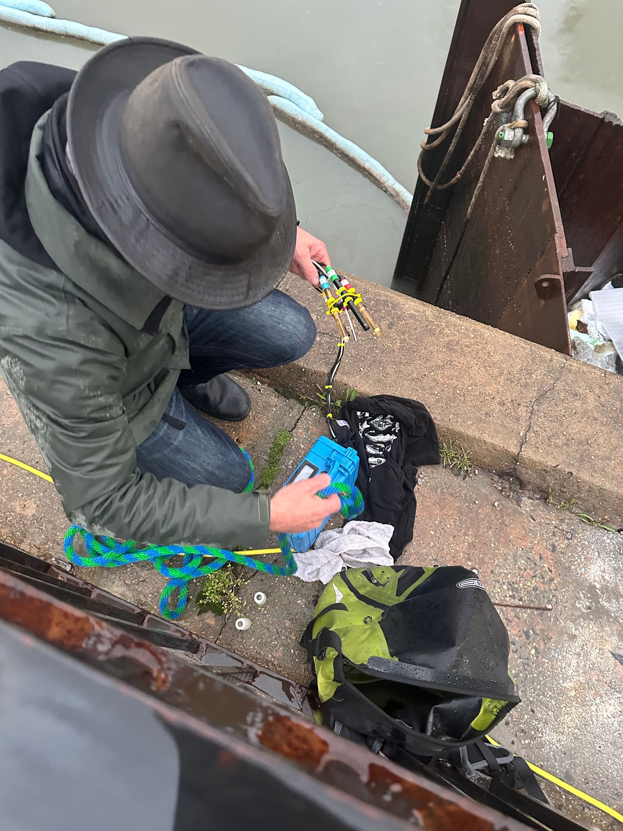 A man wearing a fedora crouches by the Gowanus Canal, holding a tangled web of multicolored ropes.