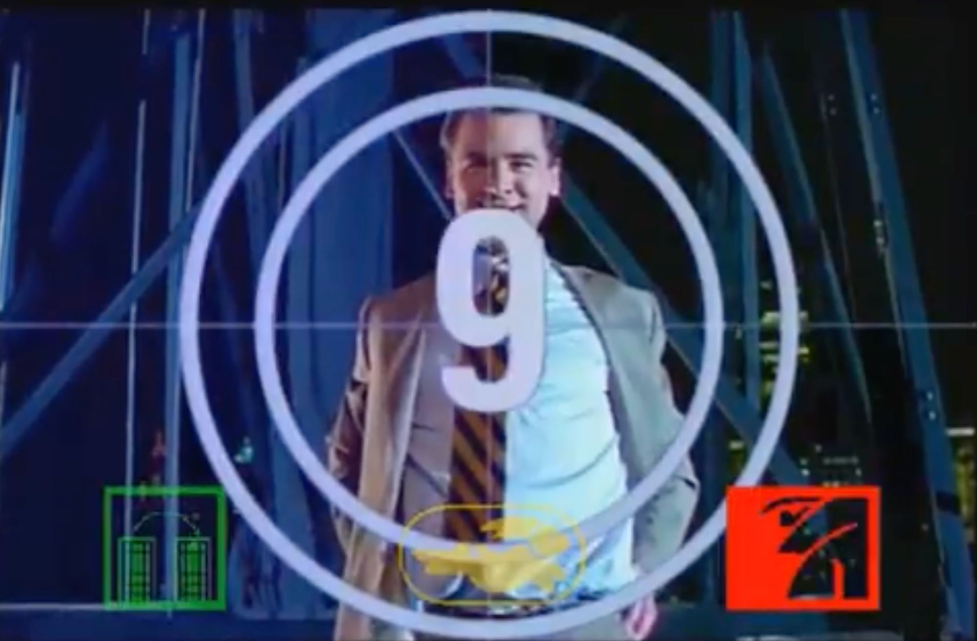 A man stands and looks expectantly at the camera in a '90s movie, with a large clock, frozen at nine seconds, partially covering his torso. 
