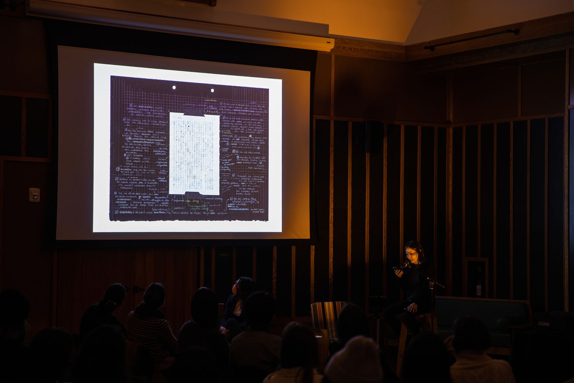 A dark photograph of the artist Asuka Goto standing at a podium and presenting her artist book, with a projection slide of one of the transcription pages of the artist book